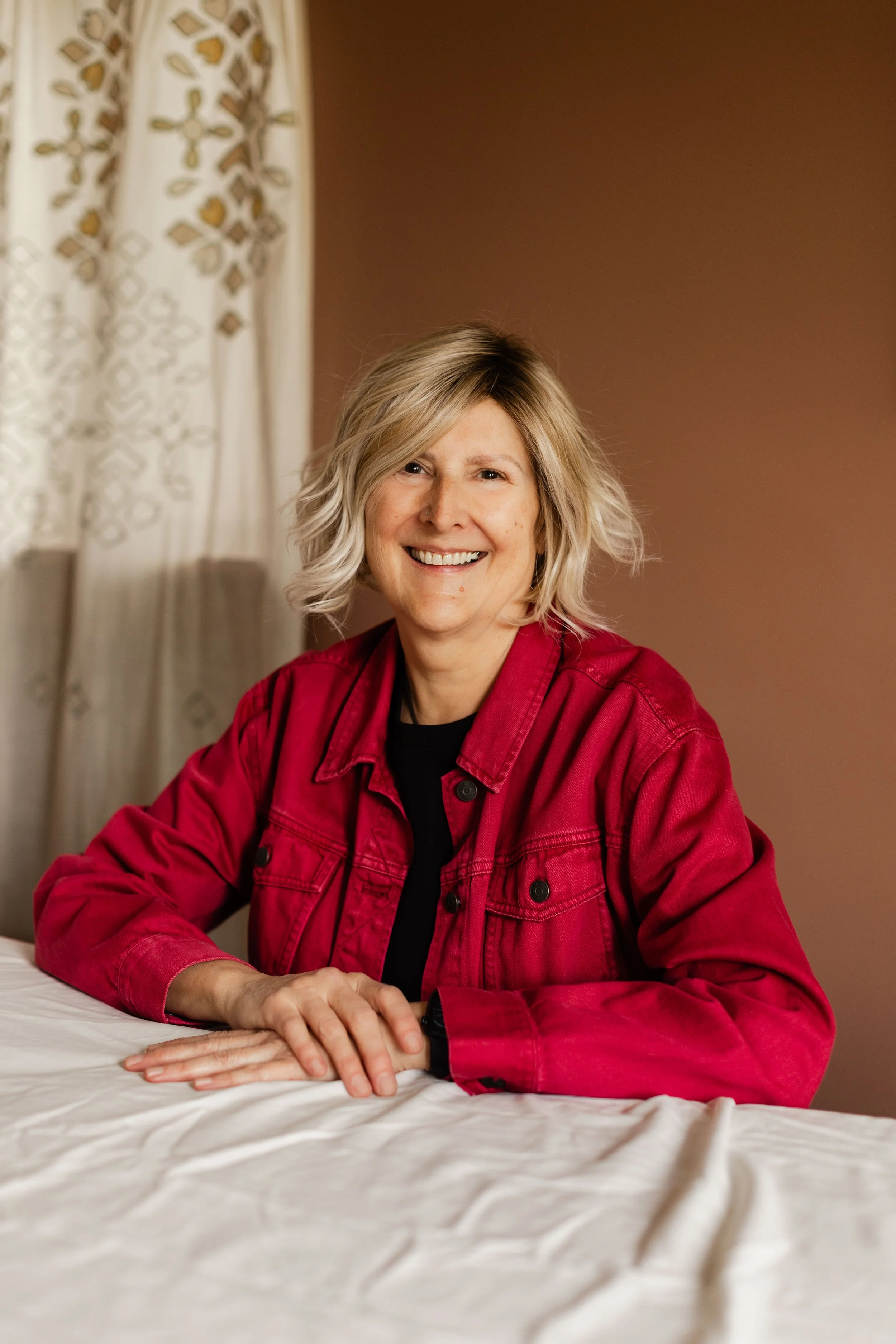 A woman with short blonde hair, smiling, wearing a red jacket and black shirt, sitting at a table with a beige tablecloth, in a room with beige walls and white curtains with gold embroidered patterns.