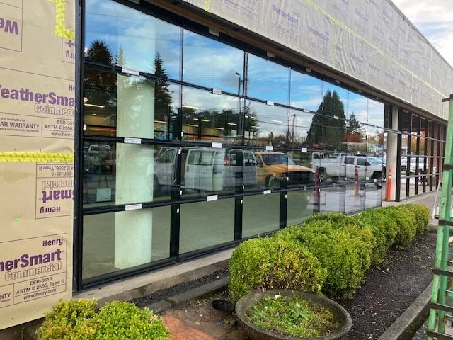 Photo of a building under construction, featuring large glass windows, construction materials, and a row of small bushes along the sidewalk.