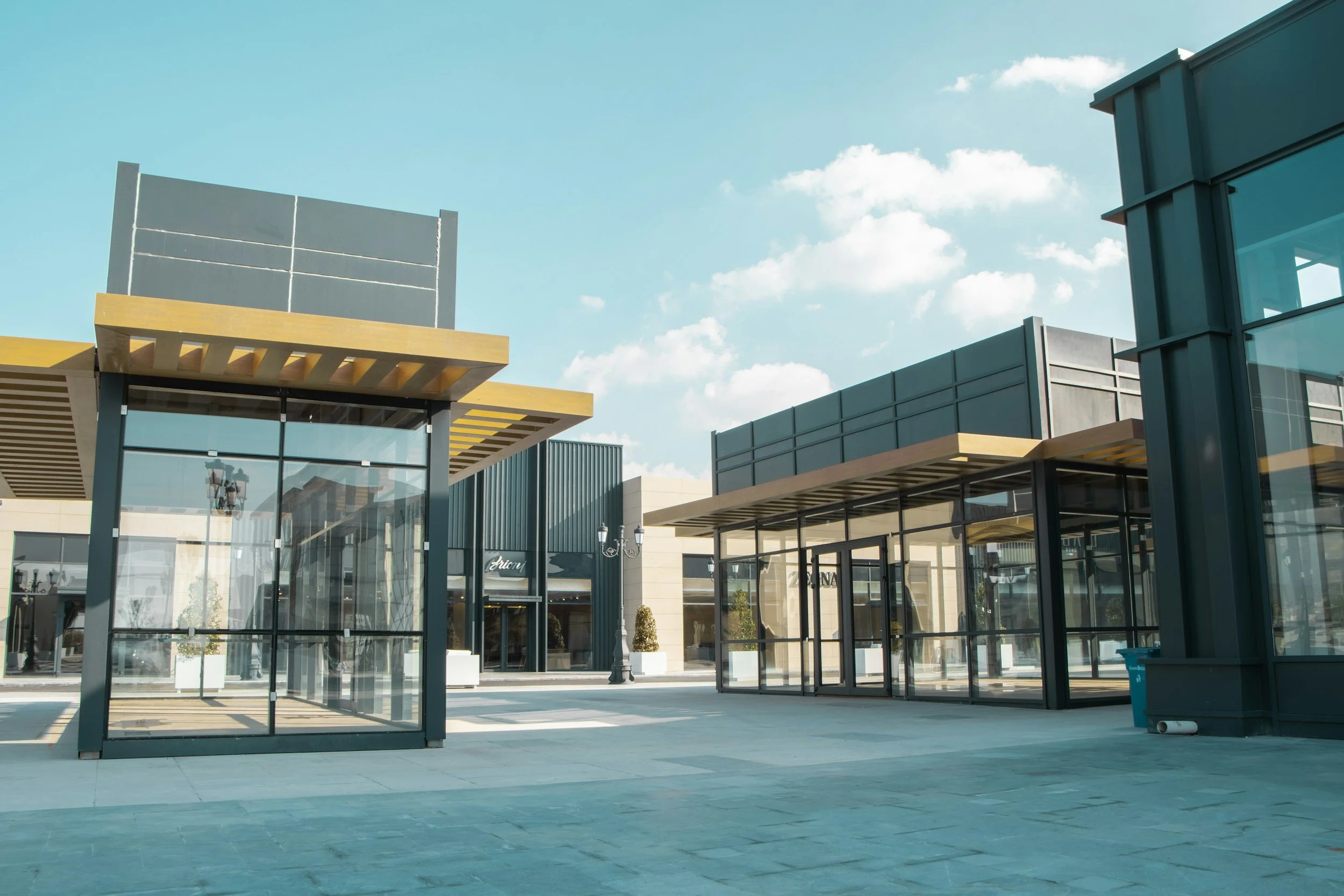 Empty outdoor shopping plaza with glass storefronts, storefronts, and a clear sky.
