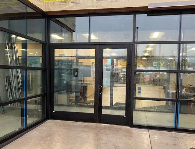 Glass double door entrance to a building with large window panes on either side, reflecting a parking lot and storefronts outside.