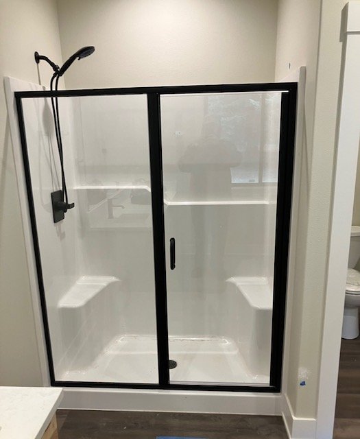 Glass shower enclosure with a black frame and sliding door, a handheld showerhead mounted on the wall, and built-in corner shelves for storage.