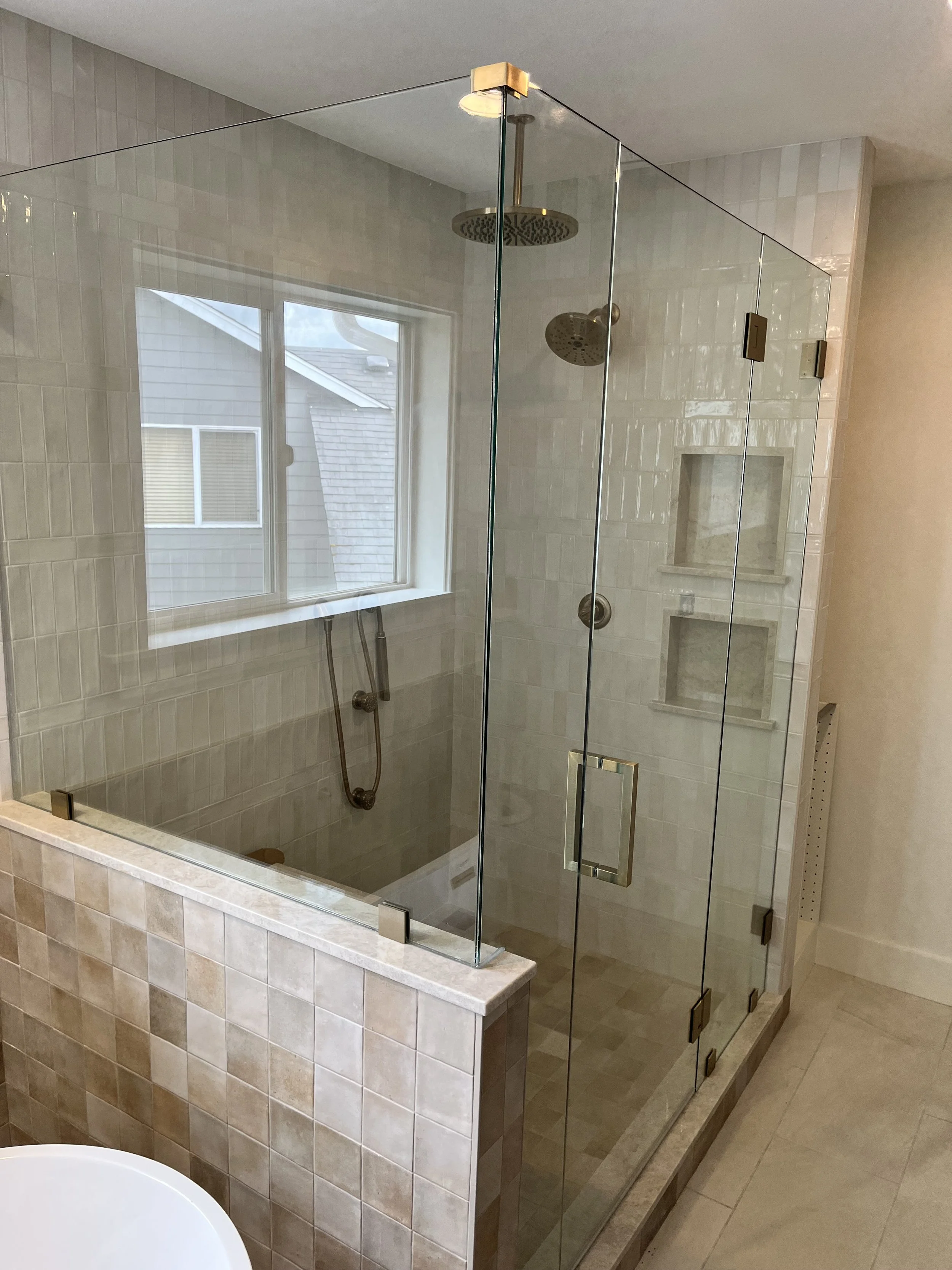 A modern bathroom shower with clear glass walls, a rainfall showerhead, a handheld showerhead, tiled walls, a built-in niche, and a window overlooking rooftops.