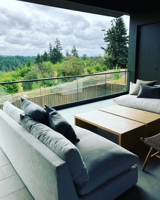 Modern living room with gray sofa and pillows, overlooking a lush green forest through a large glass window and balcony.