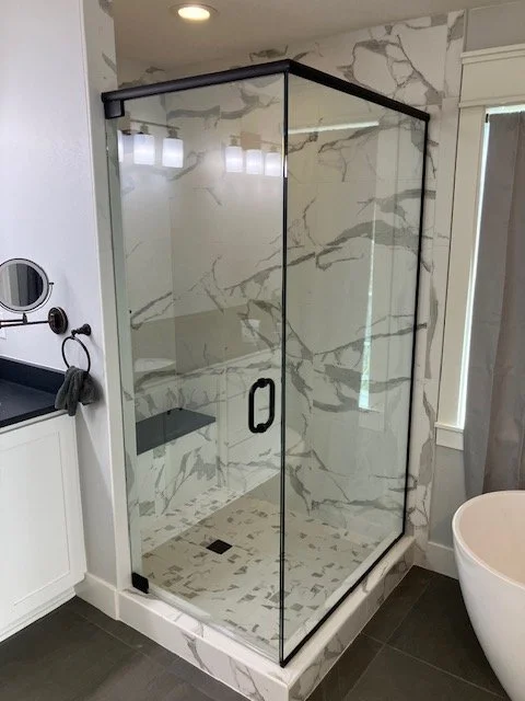 A modern walk-in shower with glass enclosure, marble walls, and a black handle, located in a bathroom with white cabinetry and a round white bathtub.