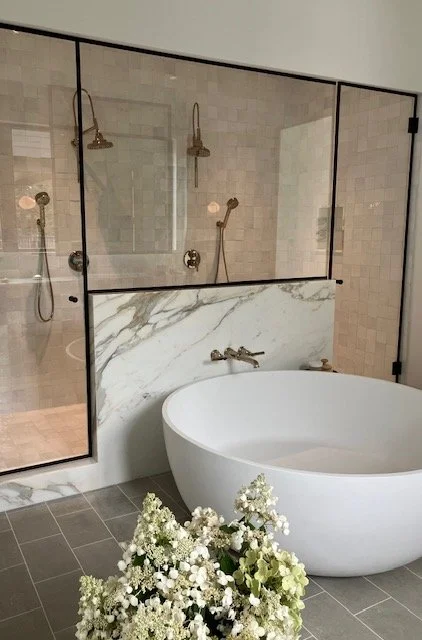 Modern bathroom with a freestanding bathtub, a separate shower area with multiple shower heads, and a glass partition, with a marble accent behind the bathtub and flowers in the foreground.