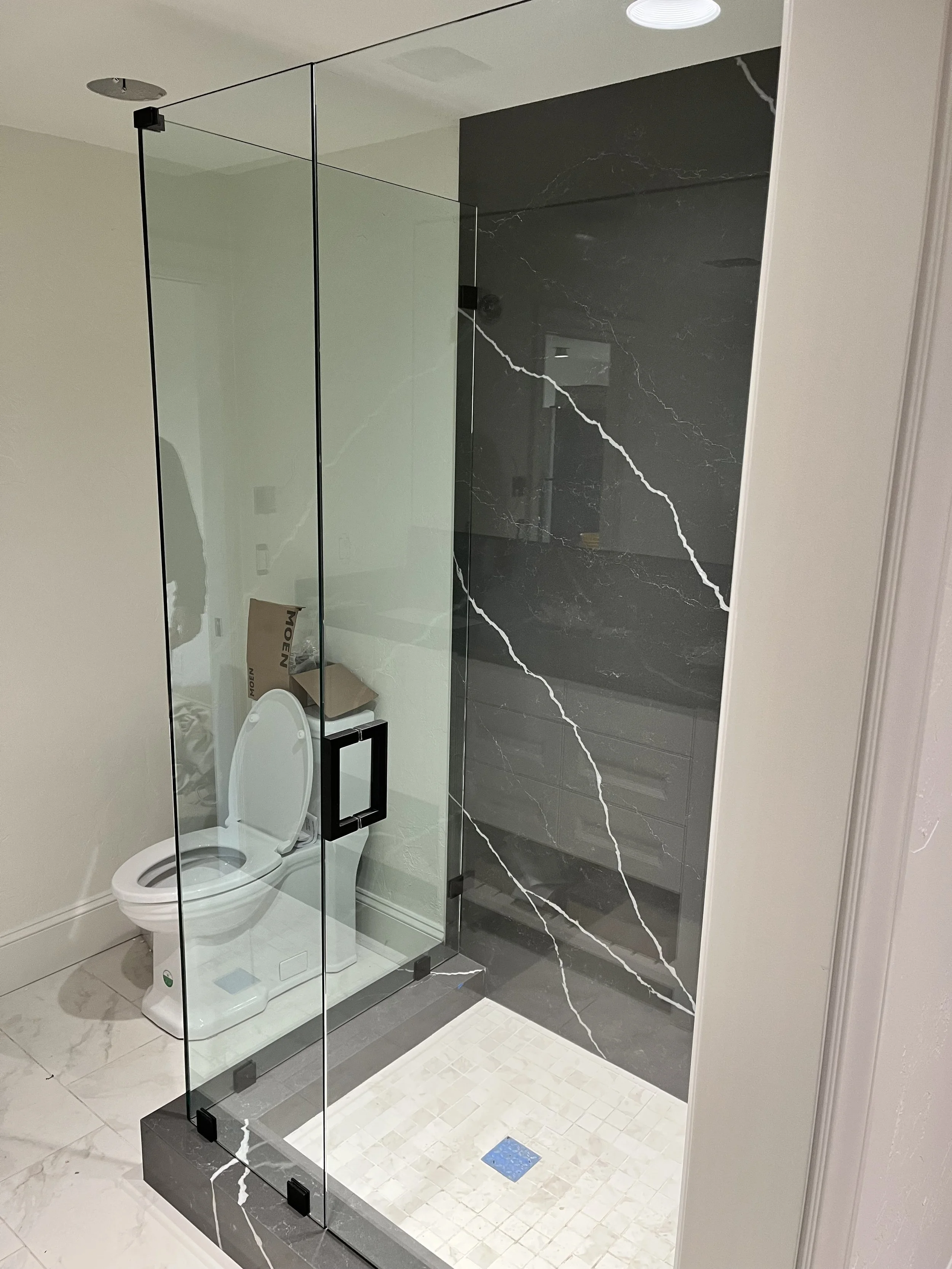 Modern bathroom with a glass shower enclosure, black marble wall with white veining, a white toilet, and beige tiled flooring.