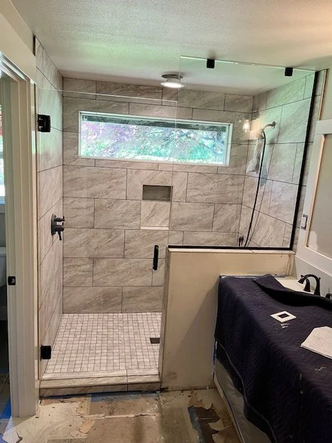 View of a partially finished bathroom shower area with beige tile walls, a small horizontal window, and a black showerhead. There is a partial wall separating the shower from the sink area, which has a black faucet and a dark countertop with various 