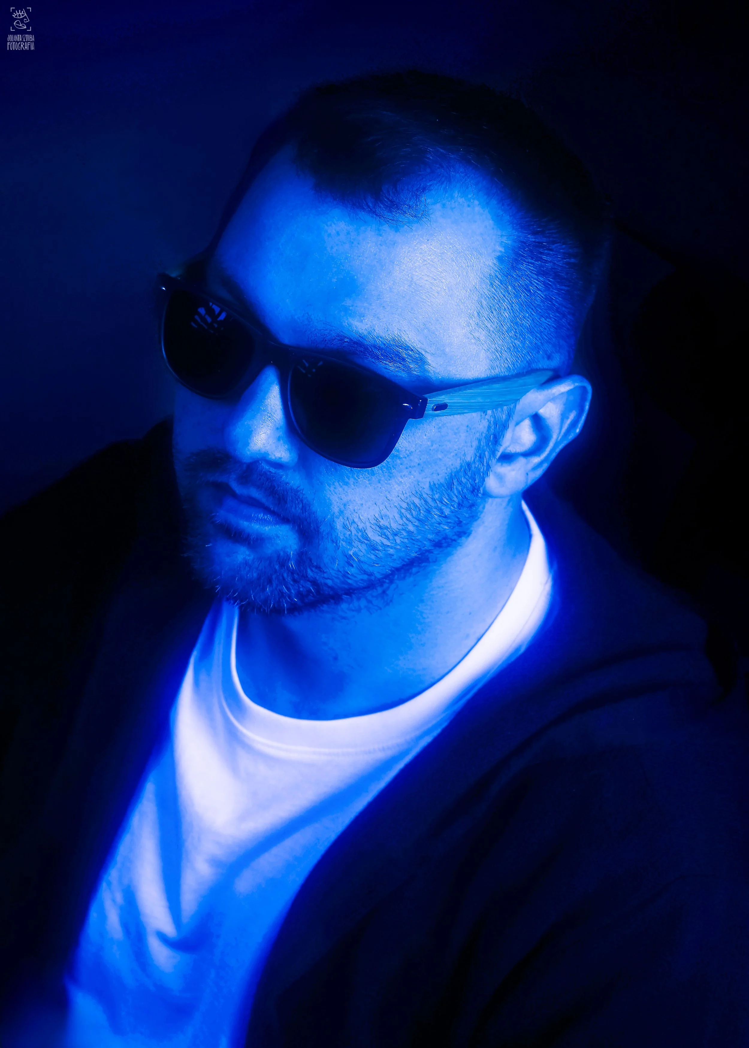 A man with short dark hair and glasses wearing a white shirt, illuminated with blue light, in a dark background.