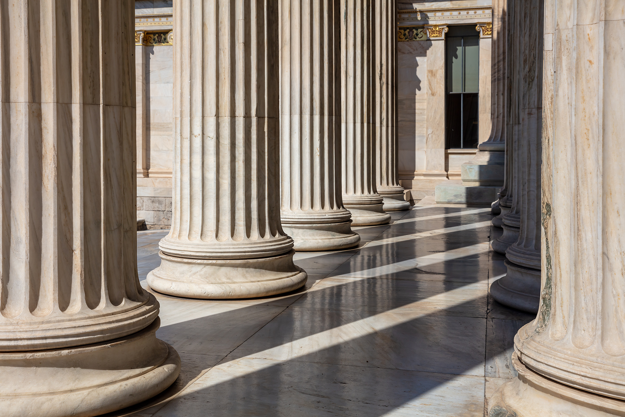 Row of large marble Greek or Roman columns with shadows cast on the floor, part of a historic building or monument.