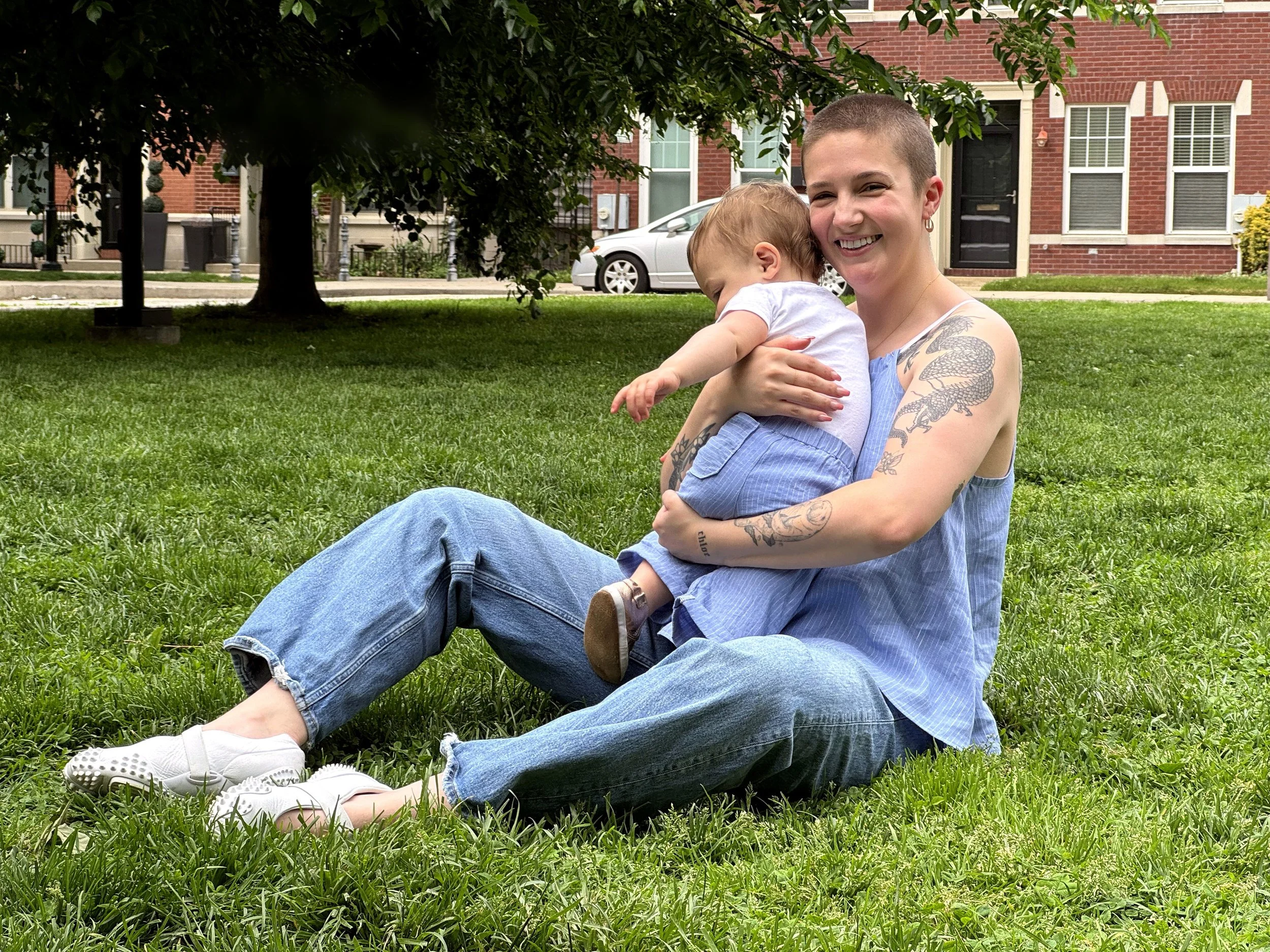 A person with short hair and tattoos on their arm sitting on the grass, holding a small child who is leaning on their shoulder, in front of a red brick building and green trees. The person and child are wearing a shirt and pants of patching fabrics.