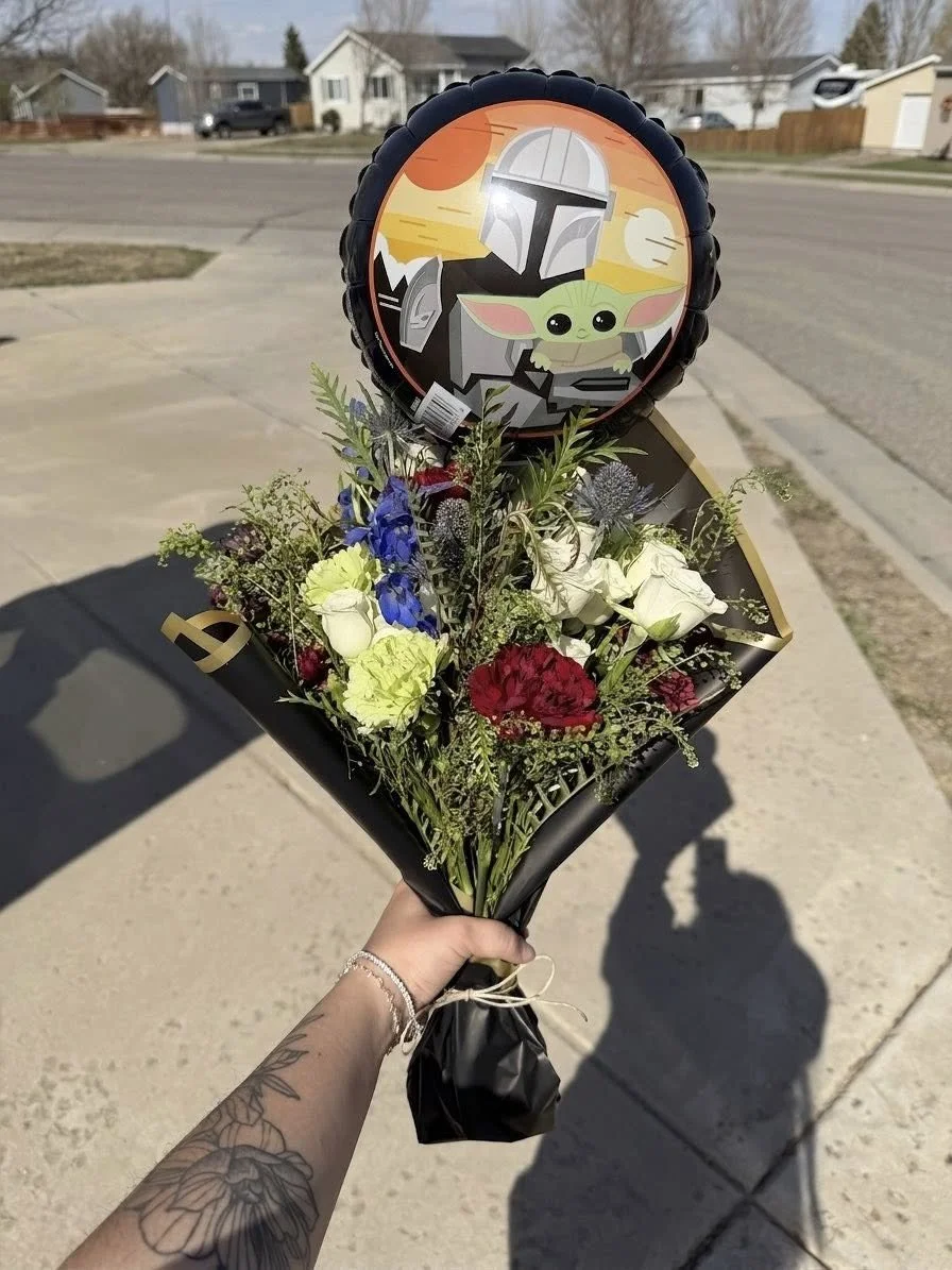A person holds a bouquet of mixed flowers, including white, yellow, red, and blue blooms, with a Baby Yoda balloon and a Baby Yoda-themed balloon of a helmeted character from Star Wars, against a residential neighborhood background.