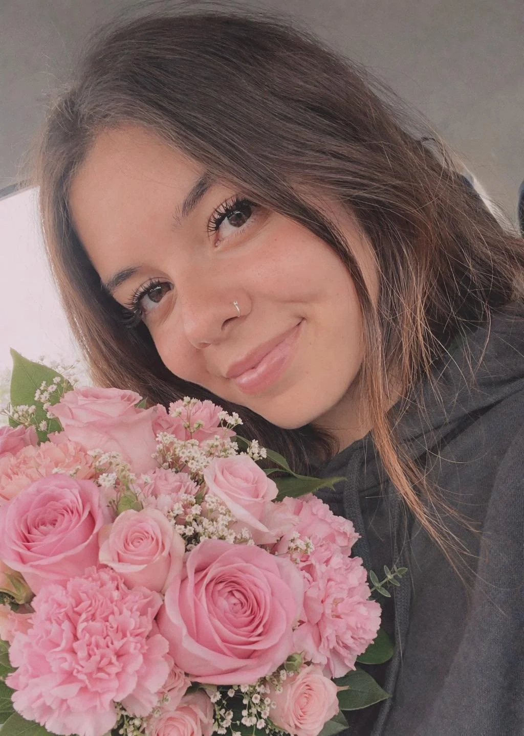 A woman with brown hair, wearing a nose ring, smiling and holding a bouquet of pink flowers, possibly roses and carnations.