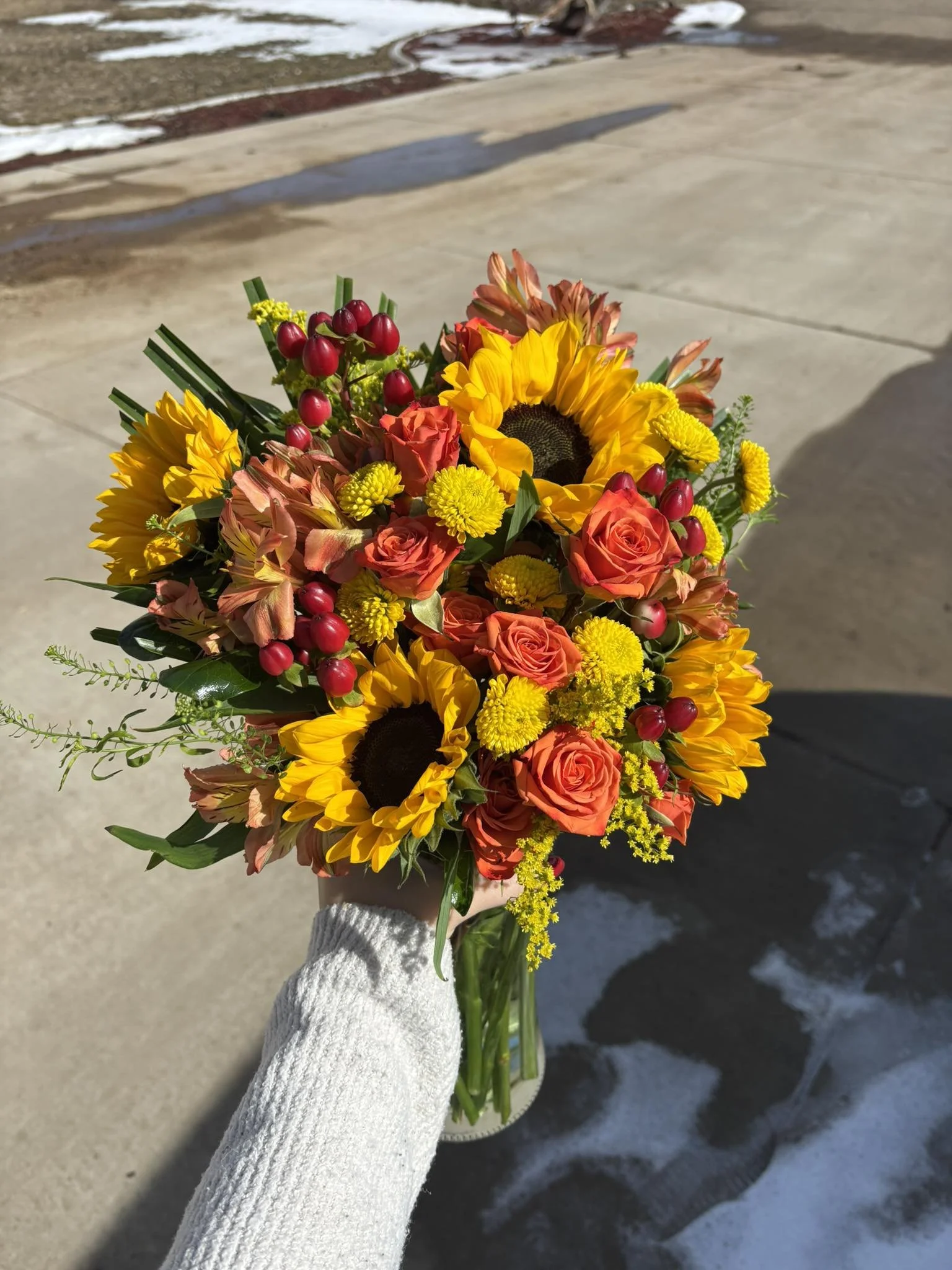 A person in a white knit sweater holds a colorful bouquet of flowers, including sunflowers, roses, and other blossoms, outdoors on a sunny day near wet concrete or pavement.