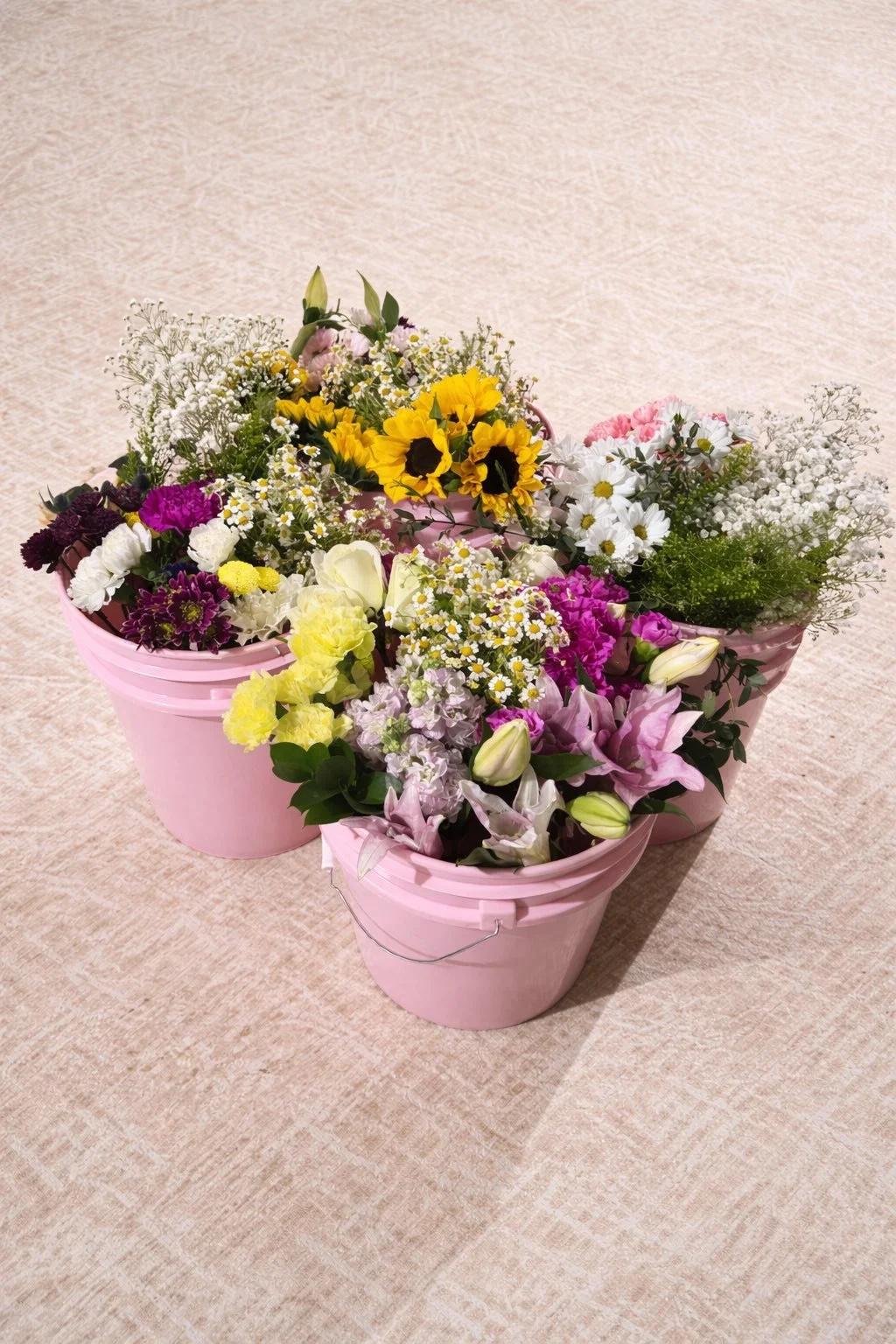 Three pink buckets filled with colorful spring flowers, including sunflowers, daisies, lilies, and baby's breath, arranged on a beige carpet.