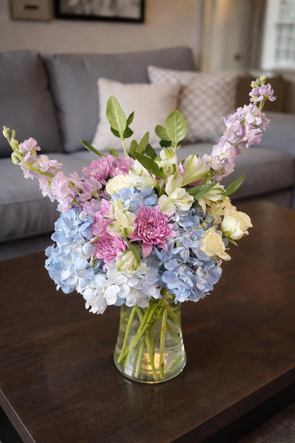 A glass vase with a bouquet of pink, blue, white, and purple flowers, placed on a dark wooden table in a living room with a gray sofa and pillows in the background.