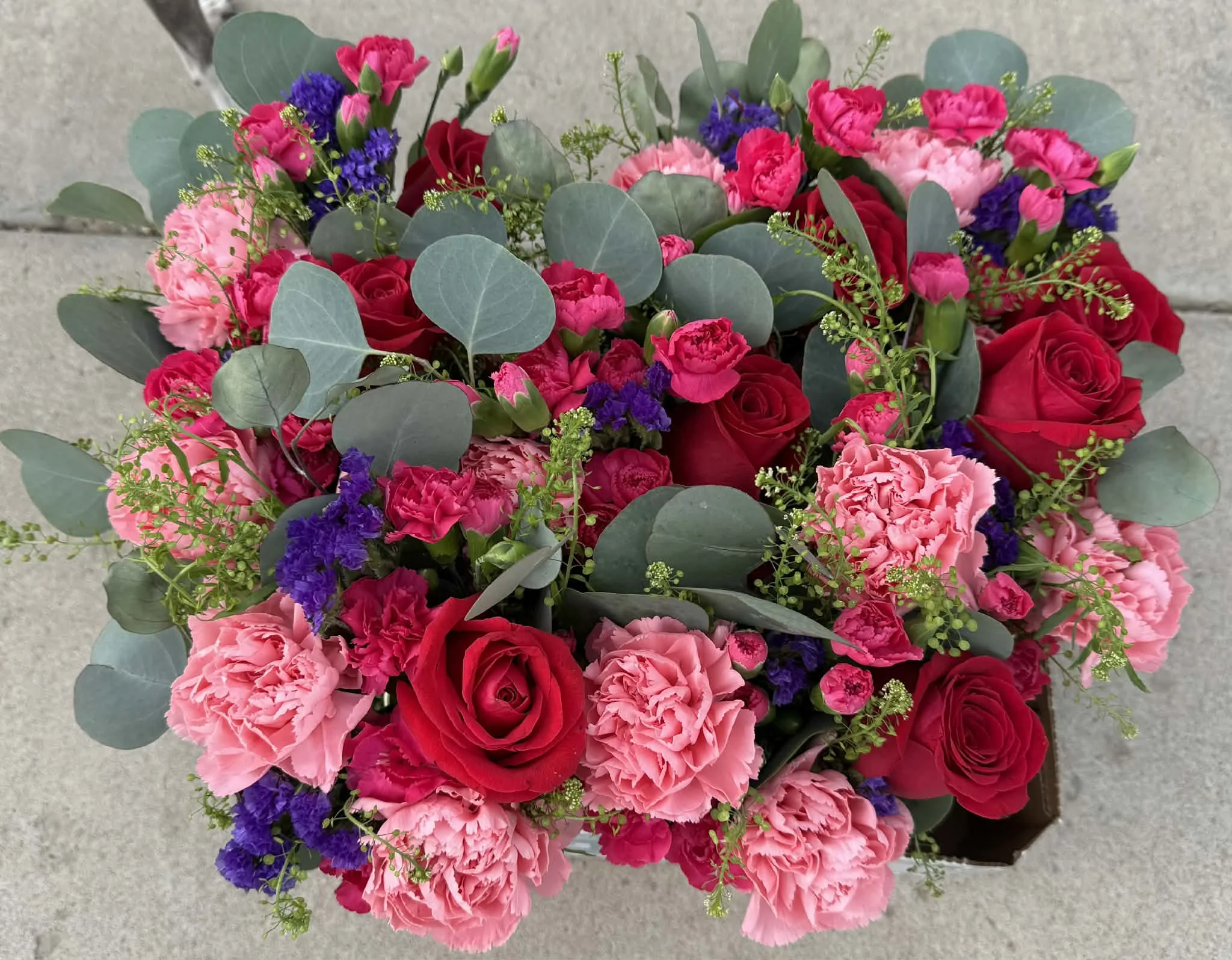 Top view of a vibrant bouquet of pink, red, purple, and peach carnations, accented with green eucalyptus leaves on a gray surface.