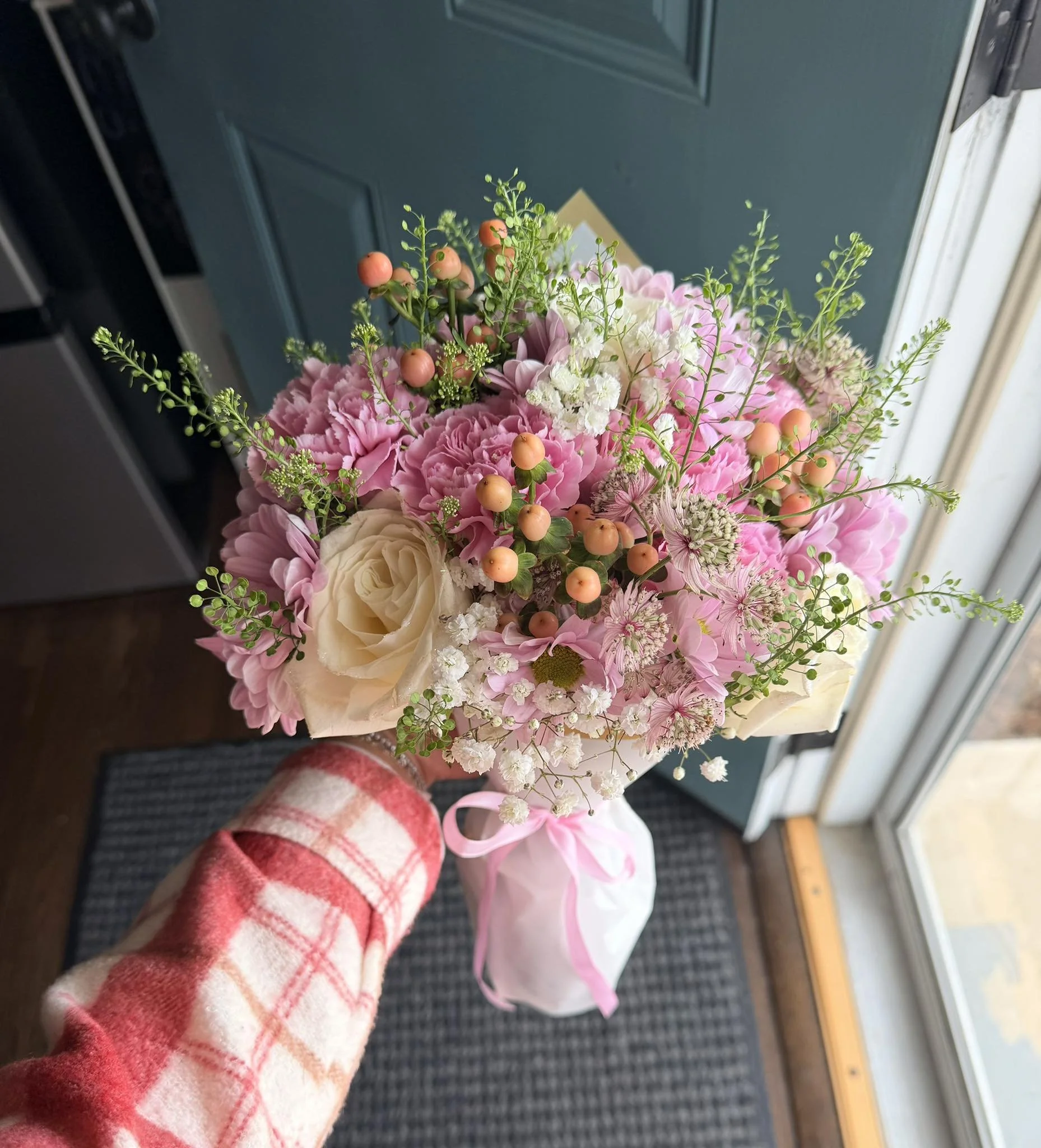 A person's hand holding a bouquet of pink and white flowers, including roses and carnations, with some small orange berries and green foliage, standing on a door mat near a blue door and a window.