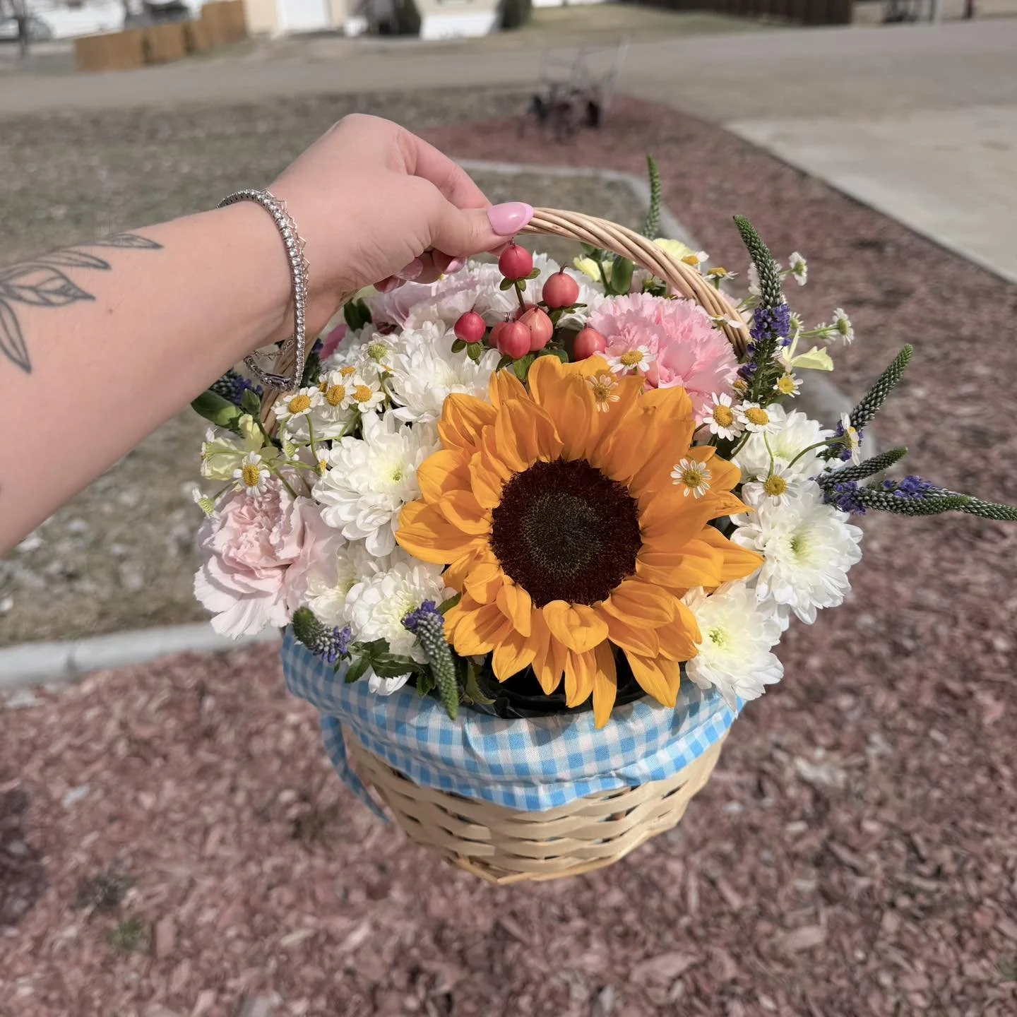 A hand holding a basket of colorful flowers with a sunflower, pink and white carnations, daisies, and lavender, against an outdoor background.