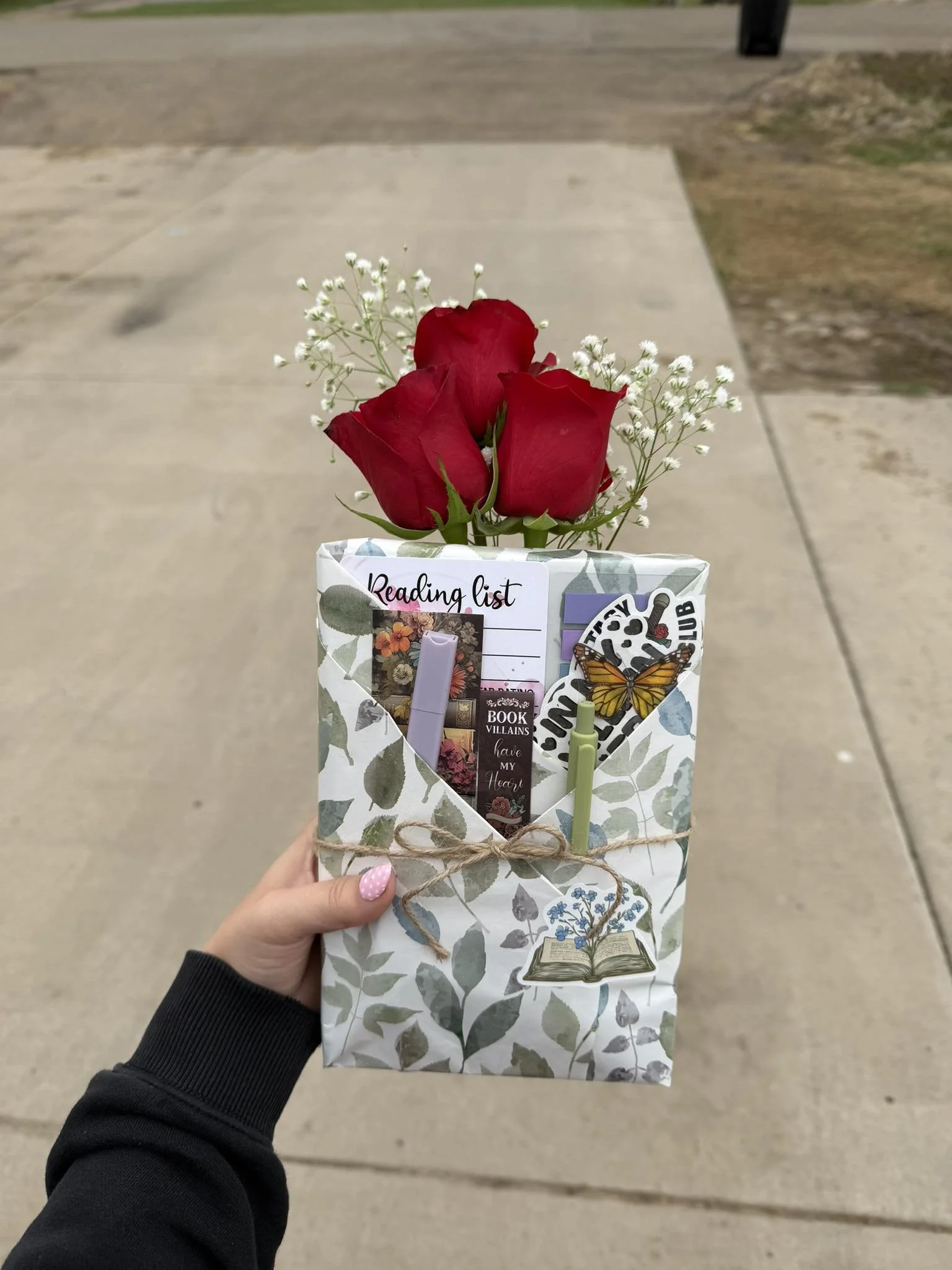 A hand holding a decorative paper bag with a floral pattern. Inside the bag are three red roses, small white flowers, a book-themed bookmark, a small lotion bottle, and other stationery items. The background shows a concrete sidewalk and some grass.