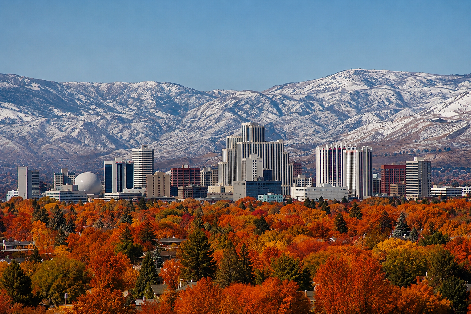 City skyline with modern buildings in front of snow-capped mountains and a foreground of vibrant autumn trees.