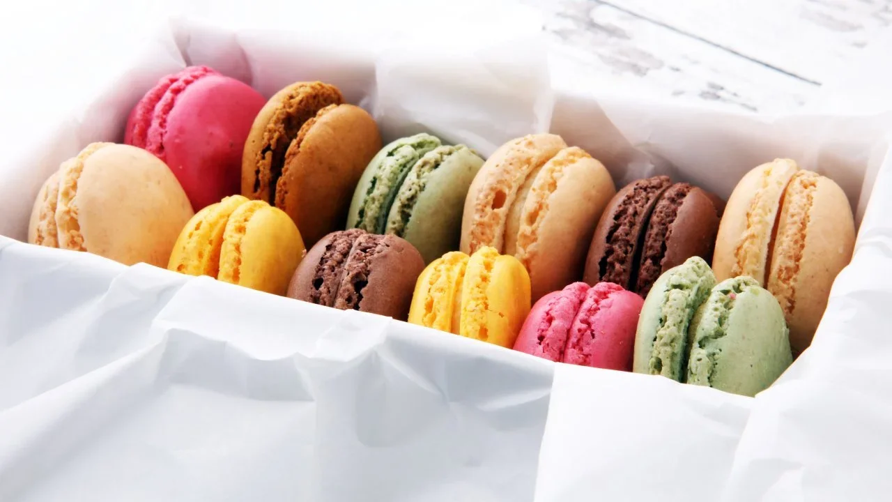 Colorful macarons in a white box, including pink, green, yellow, brown, and beige.