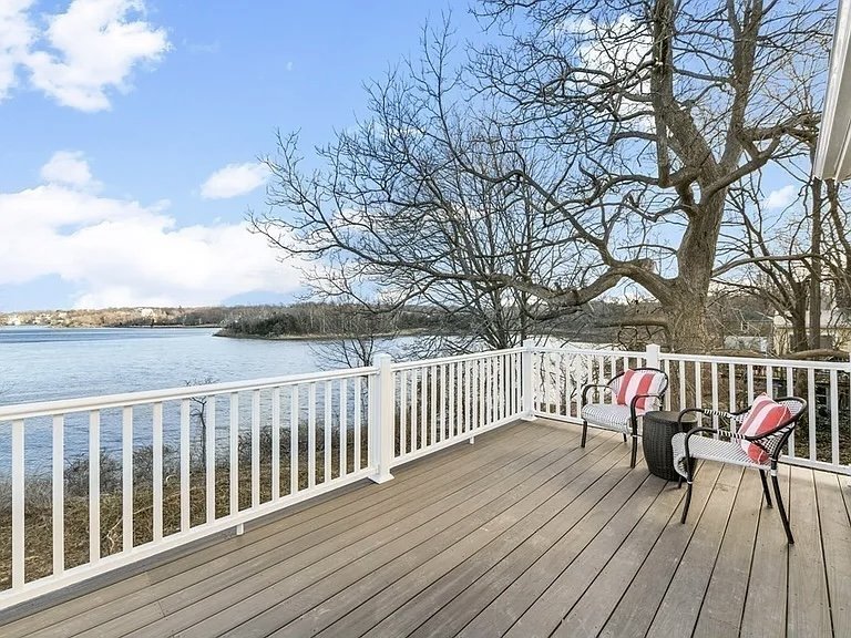 A wooden deck with white railing overlooking a river, with leafless trees and a cloudy sky in the background; two chairs with red and white striped cushions and a small black side table on the deck.