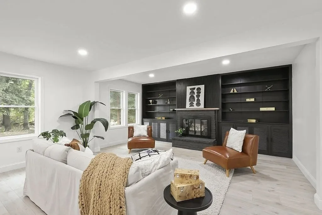 A modern living room with white walls, large windows, a white sofa with a beige blanket, two brown leather chairs, a black fireplace with built-in shelves, a framed abstract art piece, green plants, and a small black side table with stacked wooden blocks.
