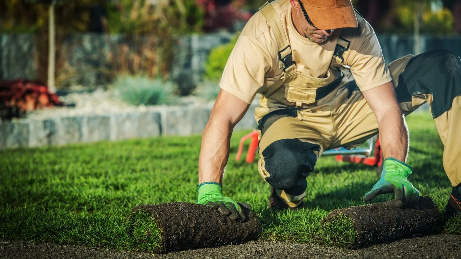Man laying on ground planting grass rolls in a yard, wearing gloves, tan work clothes, and a cap.