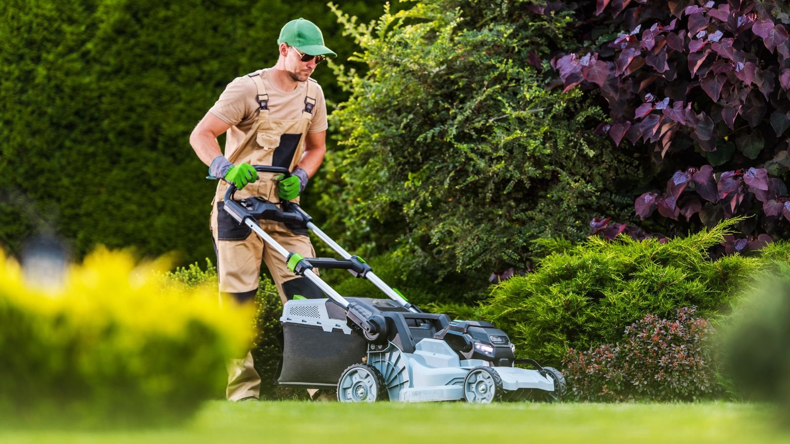 A man in outdoor clothing and green gloves mowing a lawn with a powered lawn mower in a well-maintained garden.