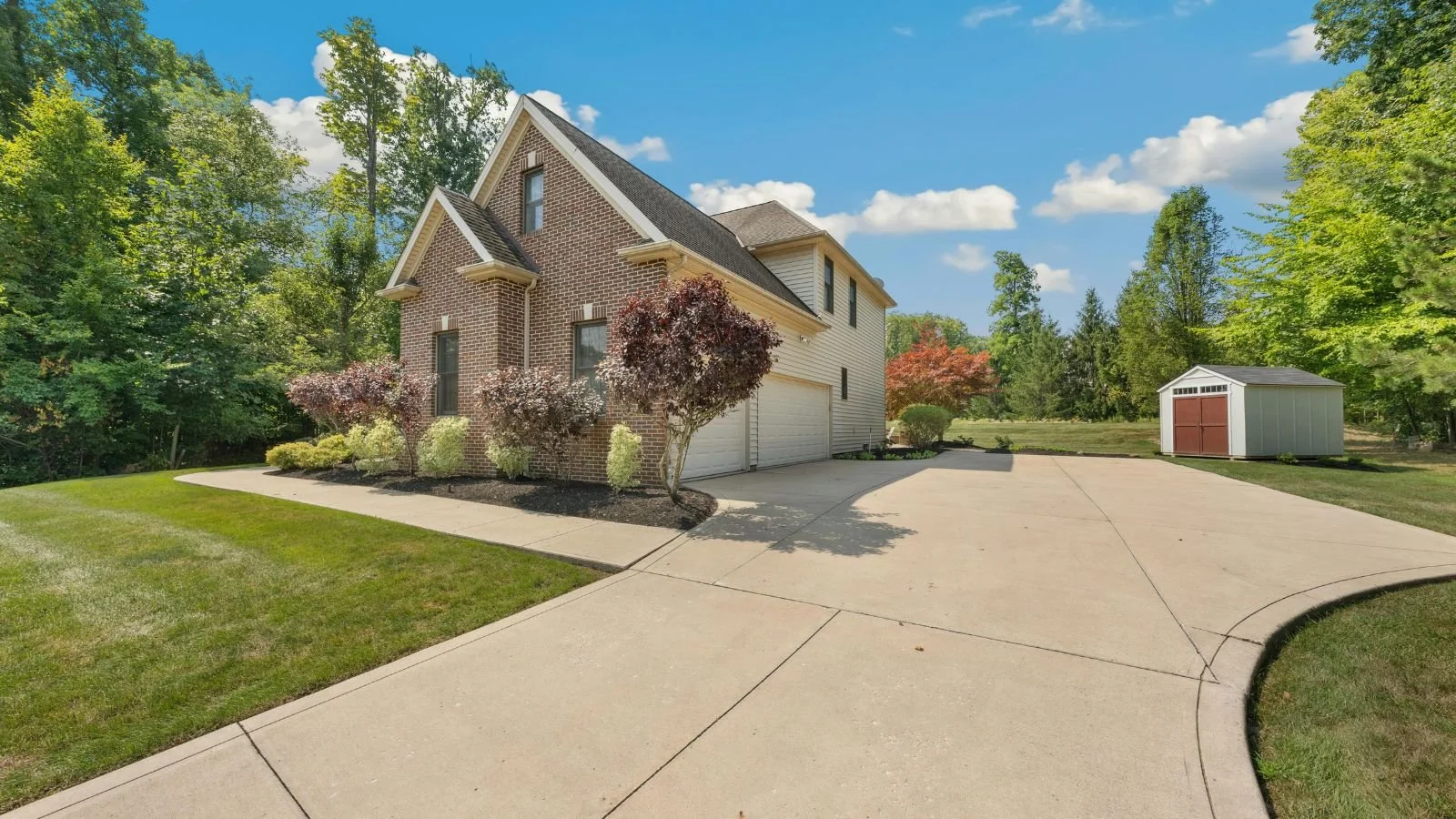 A brick house with a sloped roof and attached garage, surrounded by a well-maintained lawn, trees, and a driveway, under a partly cloudy sky.