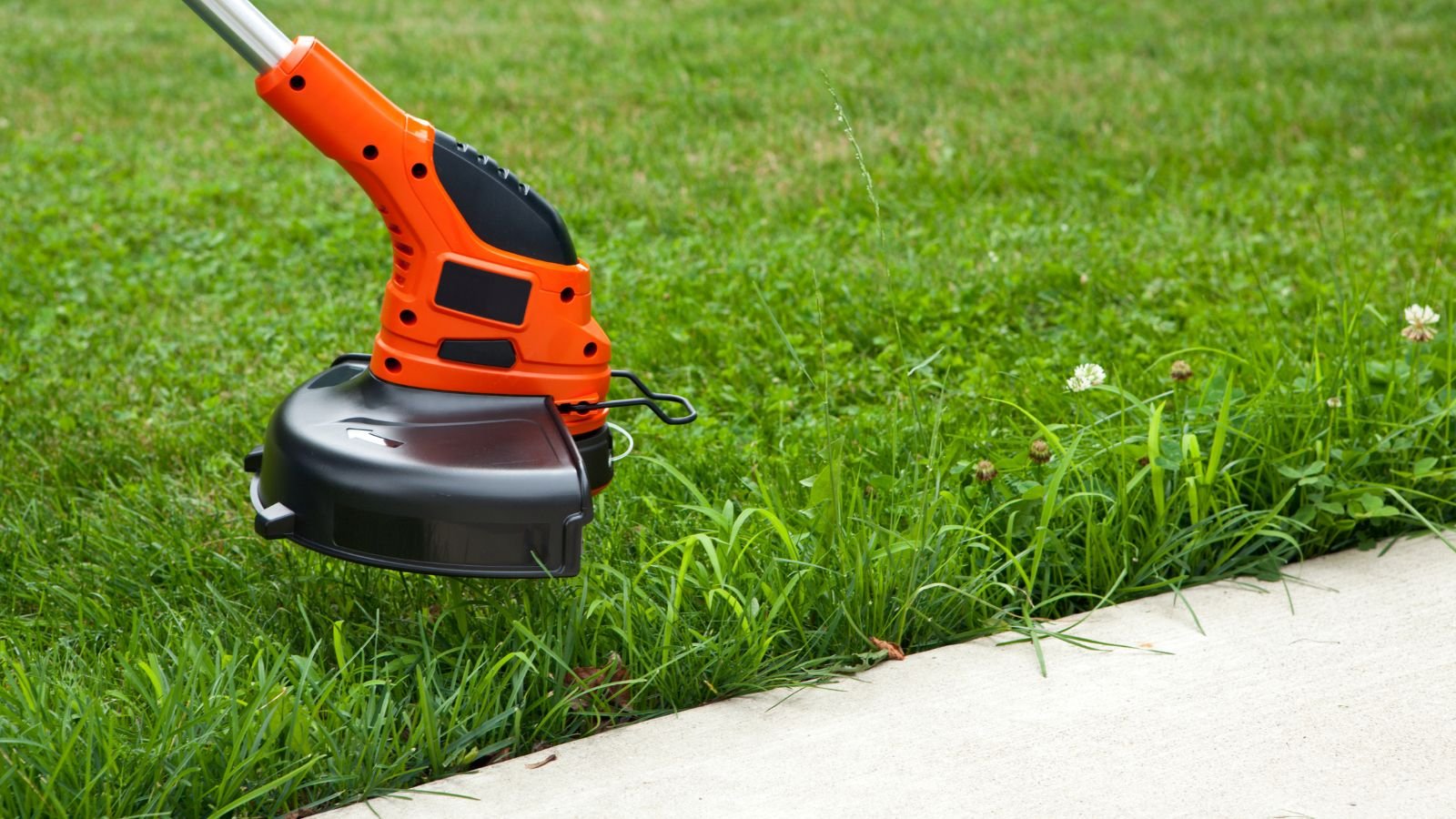 An electric weed trimmer with an orange and black handle trimming grass along the edge of a concrete sidewalk in a grassy yard.