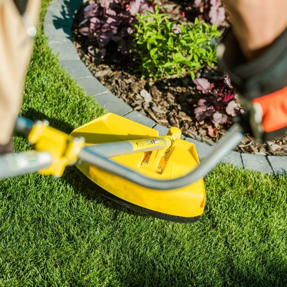 A gardener in orange and green clothing cleans a large, orange, standing lawn mower in a lush garden with green trees and bushes.