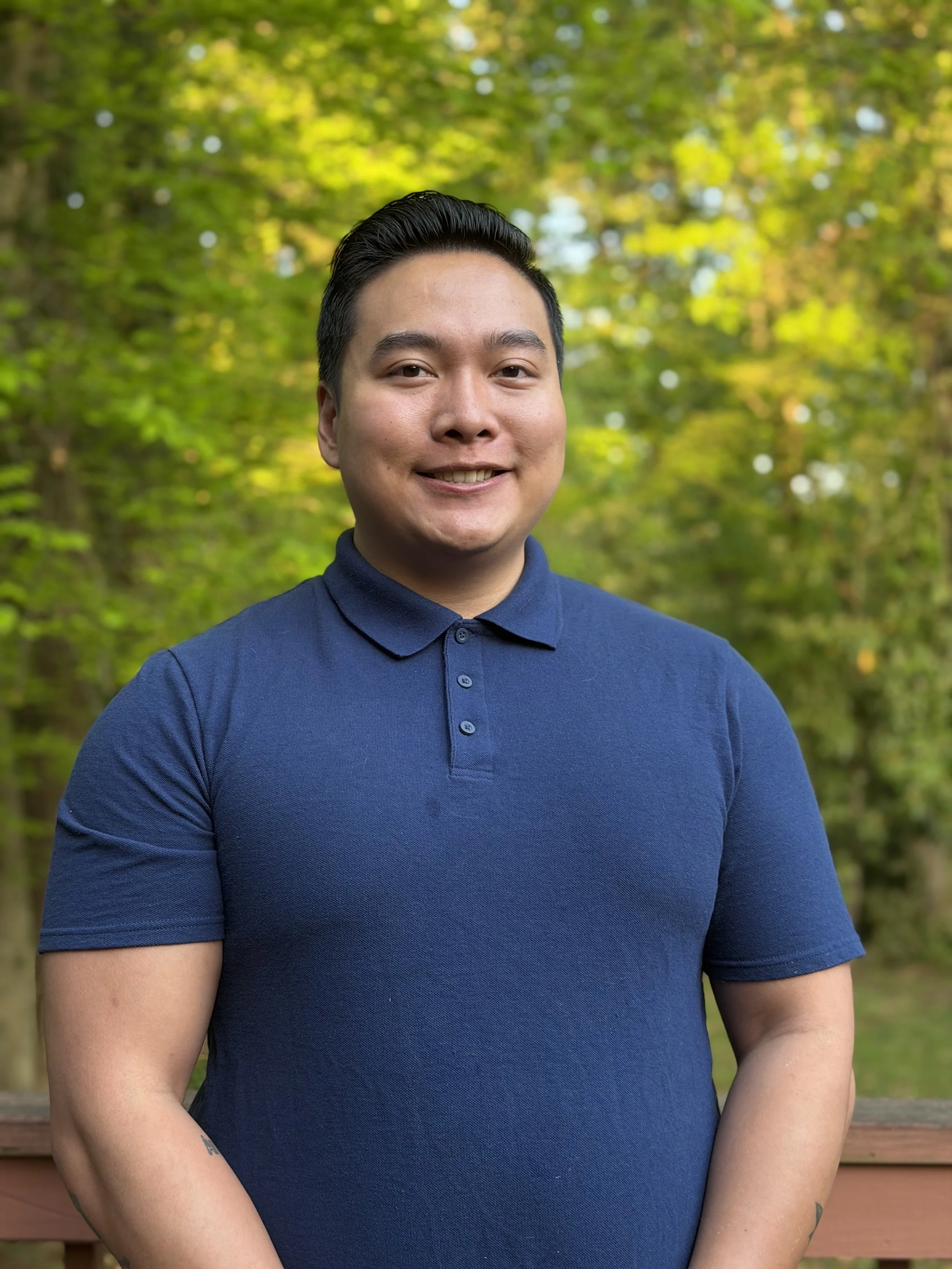 Close-up of a smiling young man with black hair, wearing a navy blue polo shirt, standing outdoors with a blurred background of green and yellow autumn foliage.