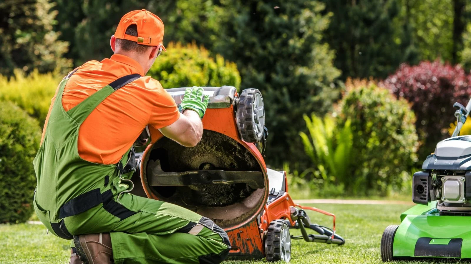 A man wearing orange and green outdoor clothing and gloves is feeding grass clippings into a paper bag shredder on a lawn with trees and shrubs in the background.