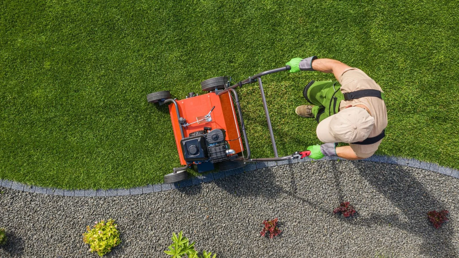 Top-down view of a person pushing a scarifier over well-maintained grass and gravel pathway, wearing a beige cap, khaki uniform, and green gloves.