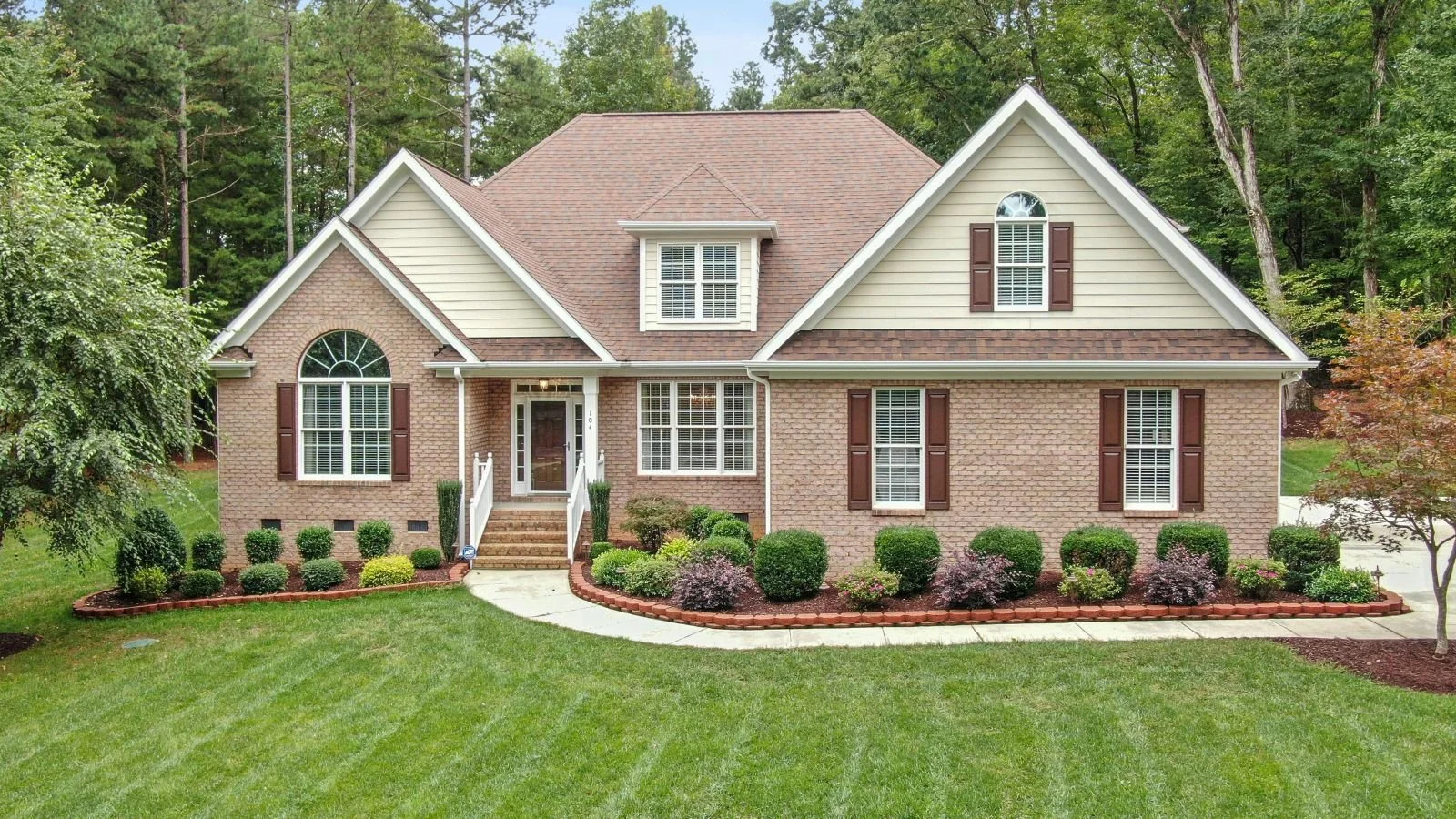 Front view of a two-story brick house with white trim, brown shutters, a covered porch, and a landscaped front yard with green grass and bushes, surrounded by trees.