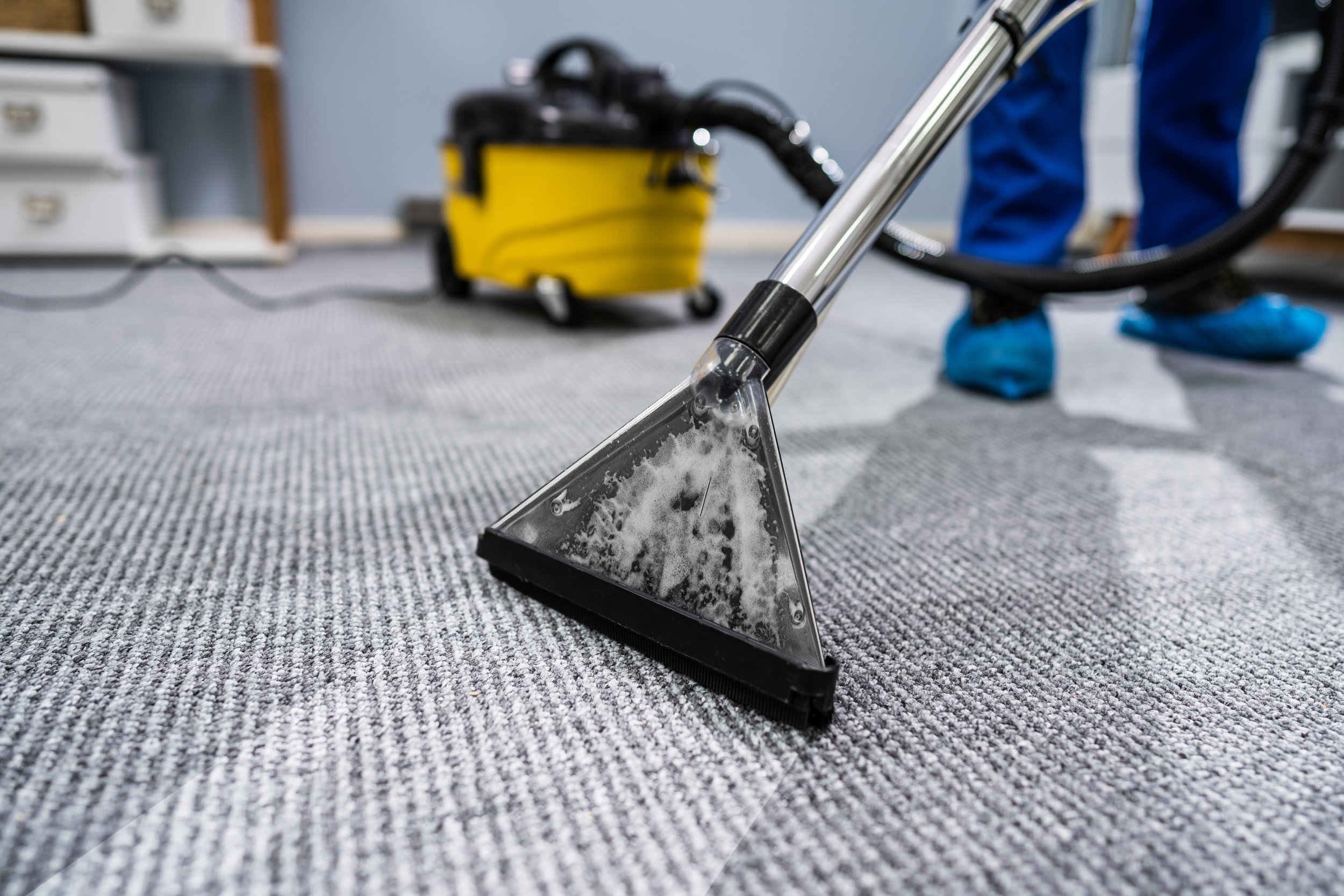 Close-up of a vacuum cleaner nozzle on a carpeted floor, with a yellow vacuum cleaner in the background.