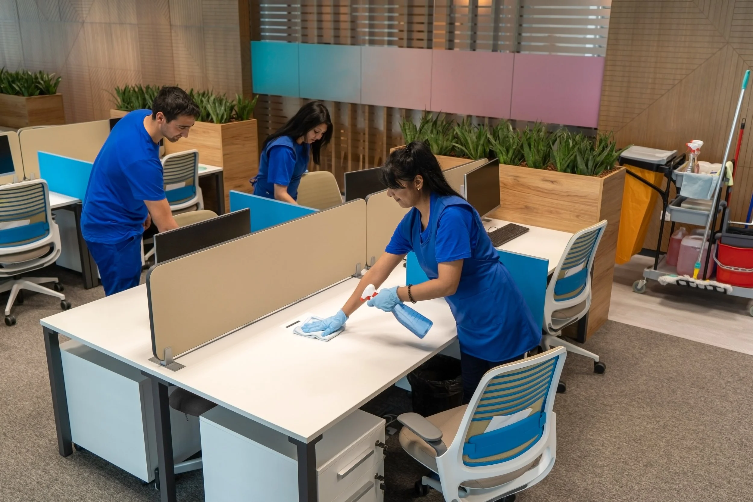 Three healthcare workers in blue scrubs cleaning and disinfecting desks in an office or waiting area, with partition screens and indoor plants in the background.