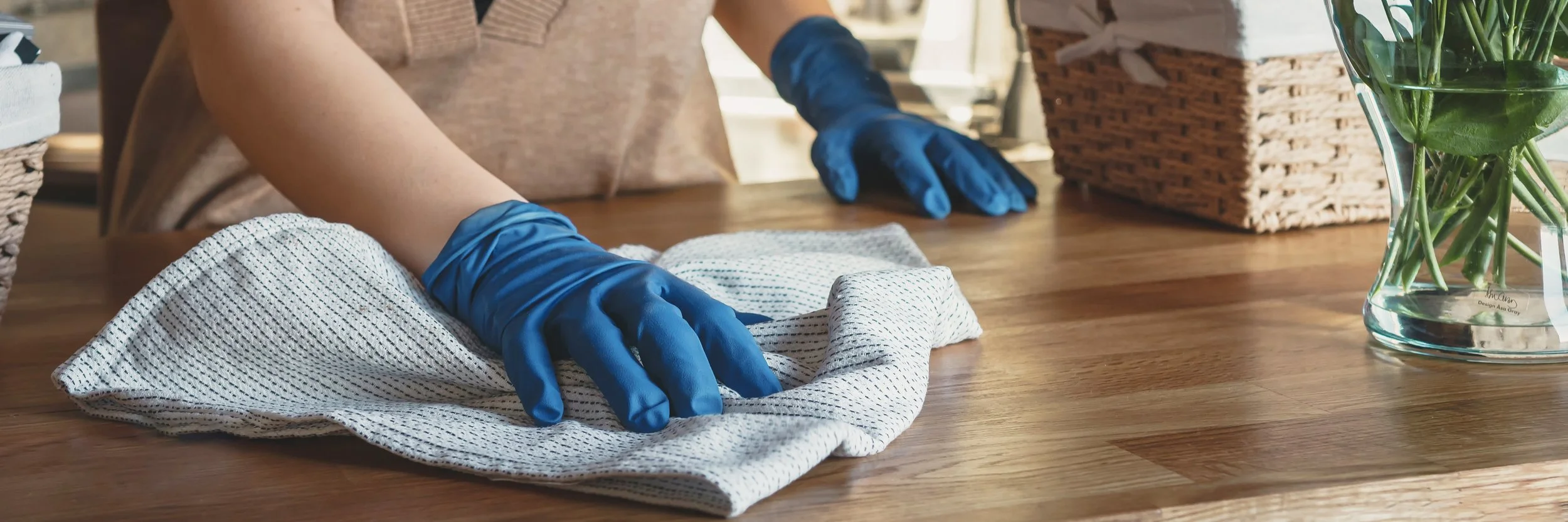 Person wearing blue gloves cleaning a wooden table with a cloth, with a glass vase of green plants in the background.