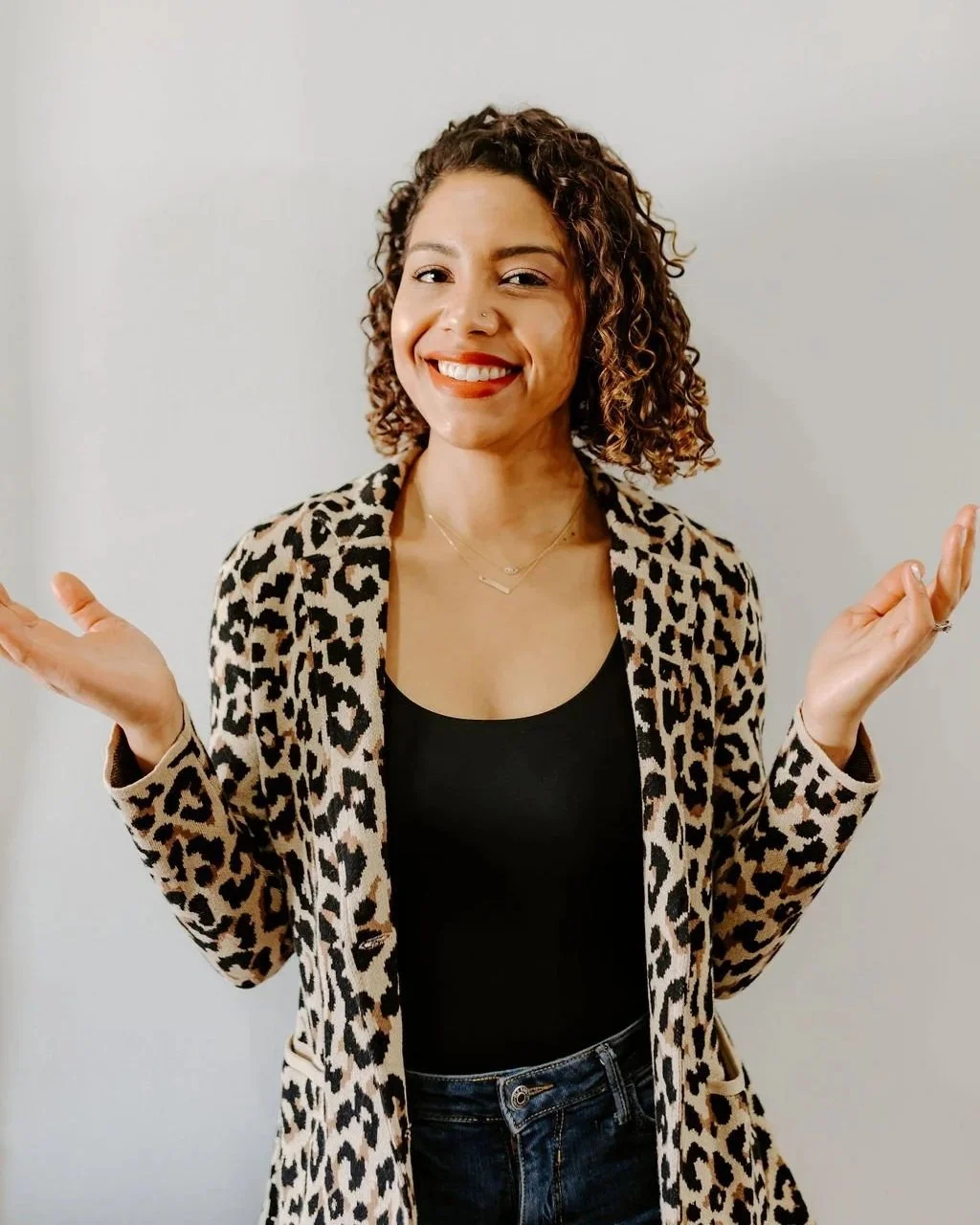 Latina woman with curly hair and red lipstick, wearing a leopard blazer holding up her hands with palms up.