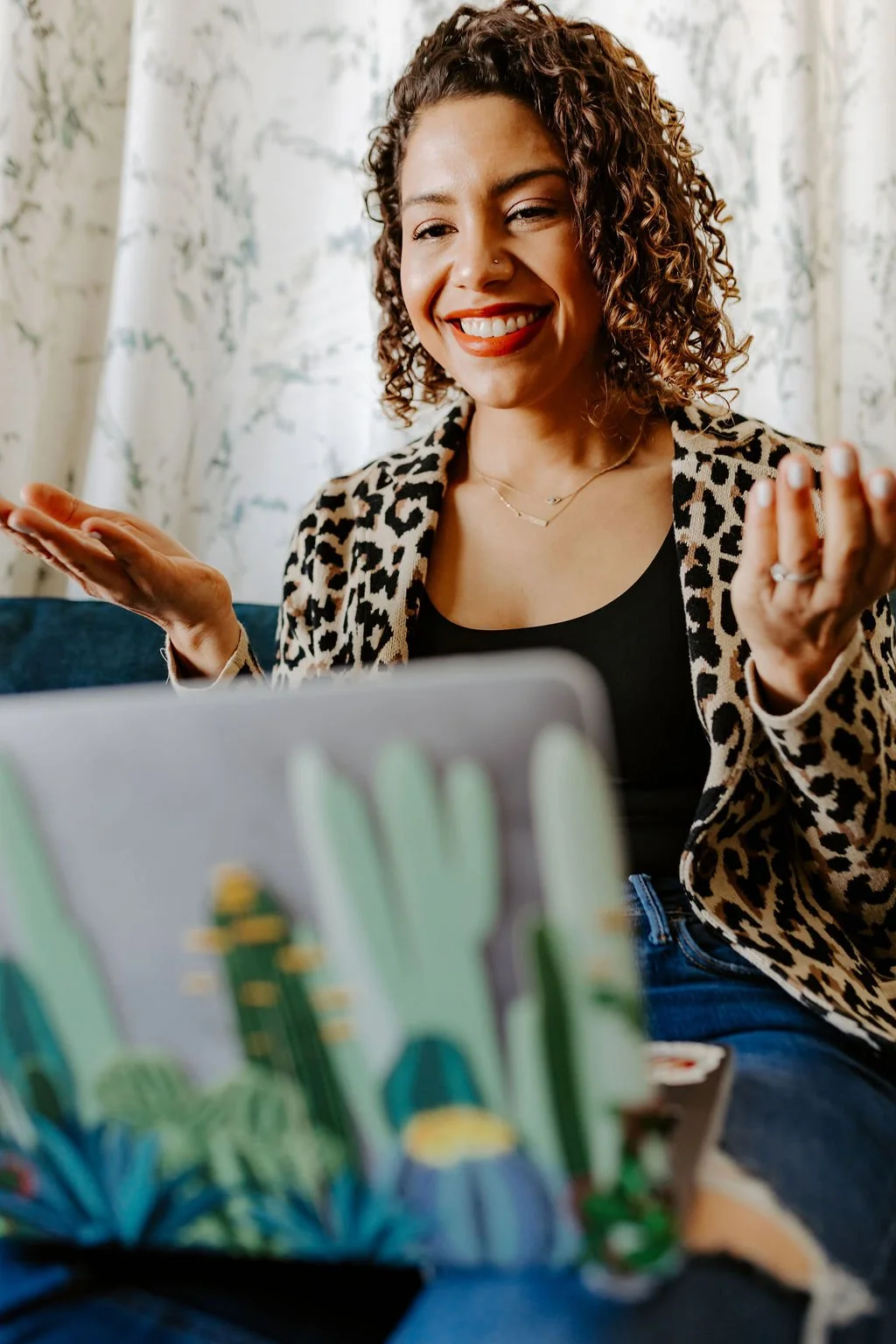 Latina woman with curly hair and red lipstick wearing a leopard blazer on a computer video call with arms up and palms facing up
