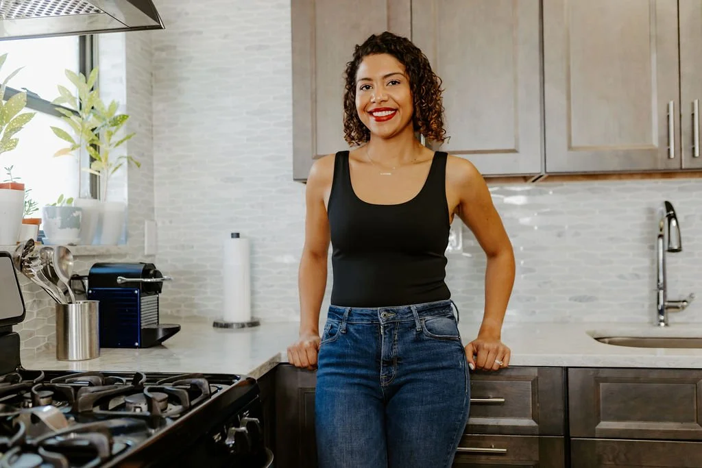 Latina woman with curly hair and red lipstick, wearing a black tank top and jeans, leaning against a kitchen counter.