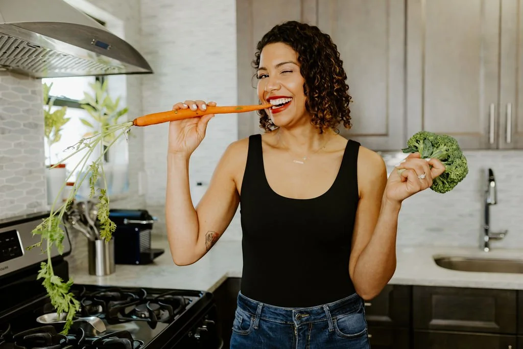 Latina women with curly hair and red lipstick, wearing a black tank top and jeans, holding a broccoli in her left hand a holding a carrot with her right hand which she is bitting.