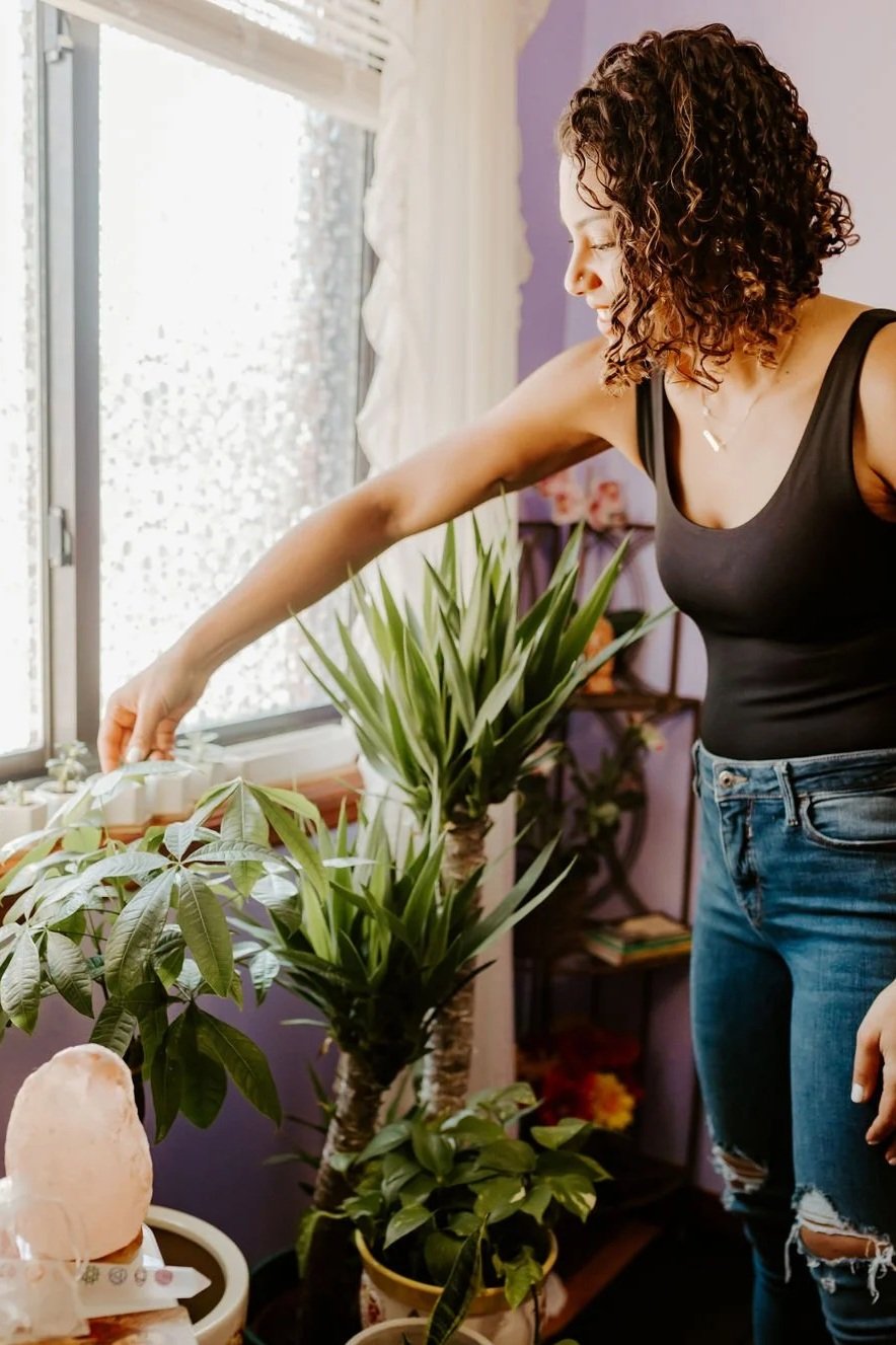 Woman with curly hair wearing a black tank top and jeans caring for plants.