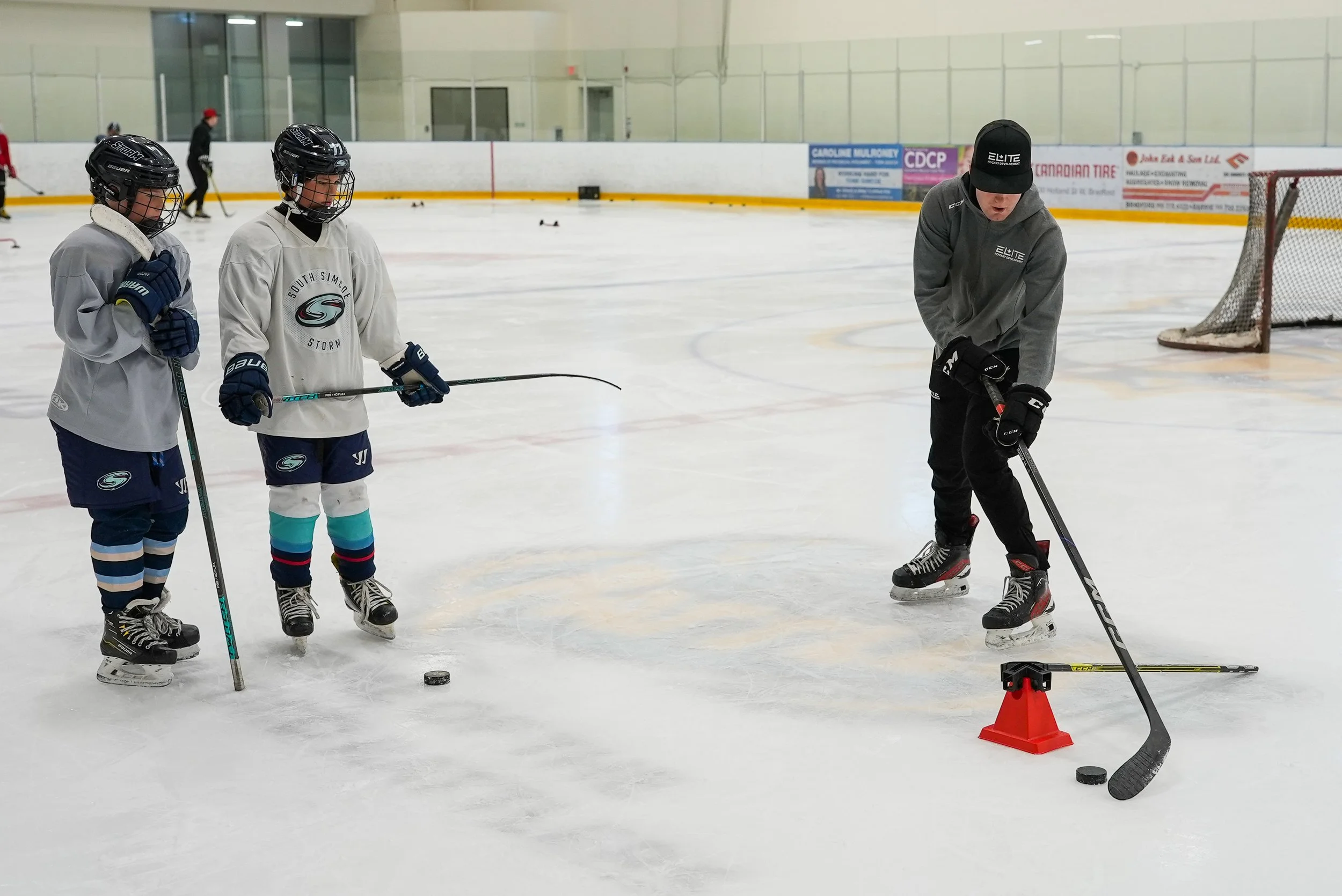 Two young hockey players wearing helmets, gloves, and jerseys listening to a coach on an indoor ice rink. The coach is demonstrating a technique near a hockey puck, with a small cone and training equipment nearby. Other players and coaches can be seen in the background.
