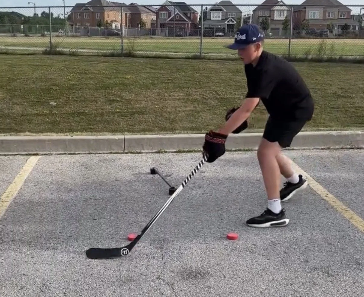 A young boy playing street hockey, wearing a blue cap, black shirt, and black shorts, using a hockey stick to hit a puck on an asphalt parking lot with yellow lines, near a grassy area and residential houses in the background.