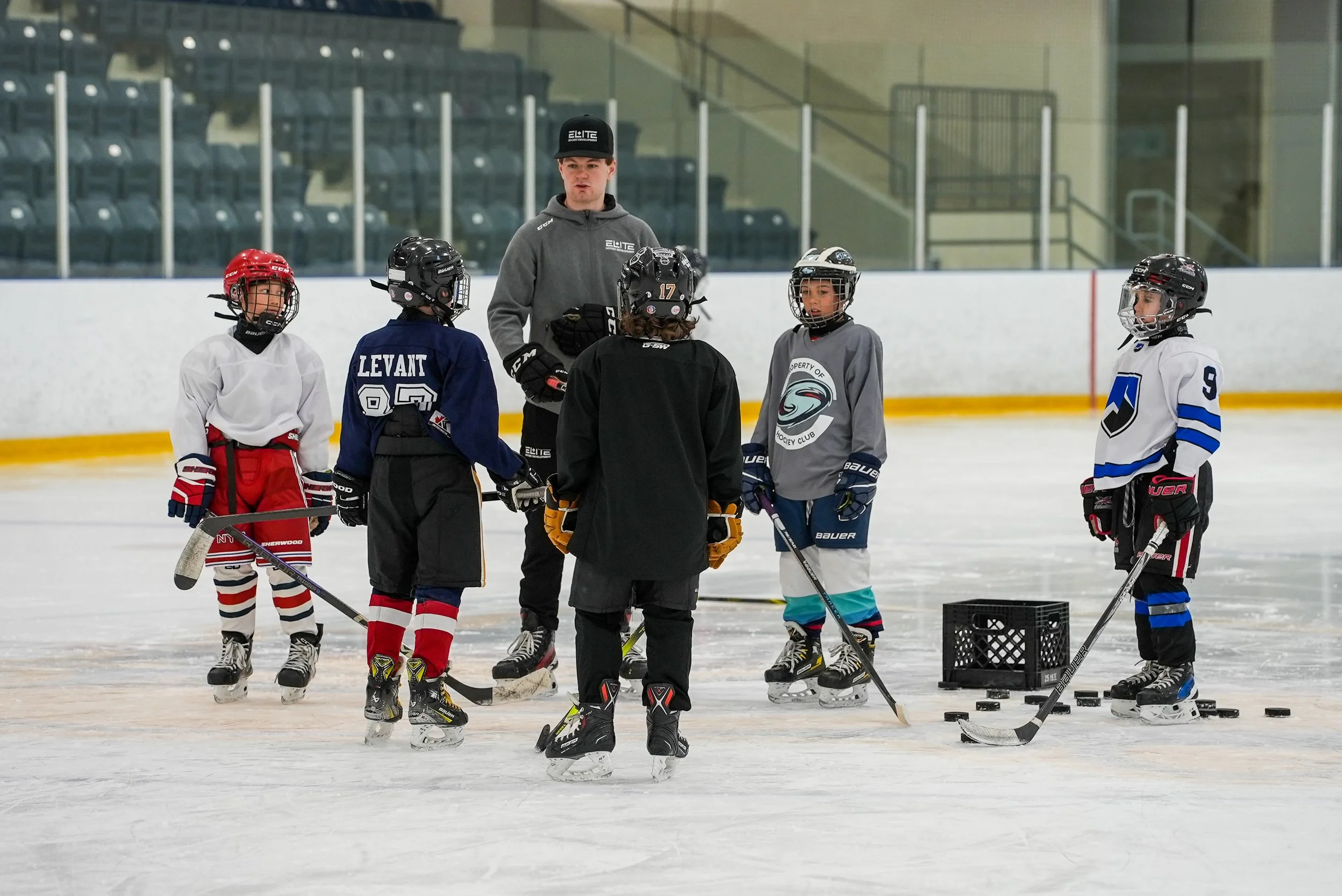 A hockey coach speaking to young players on an ice rink during practice. The children are in hockey gear, holding hockey sticks, and listening attentively.