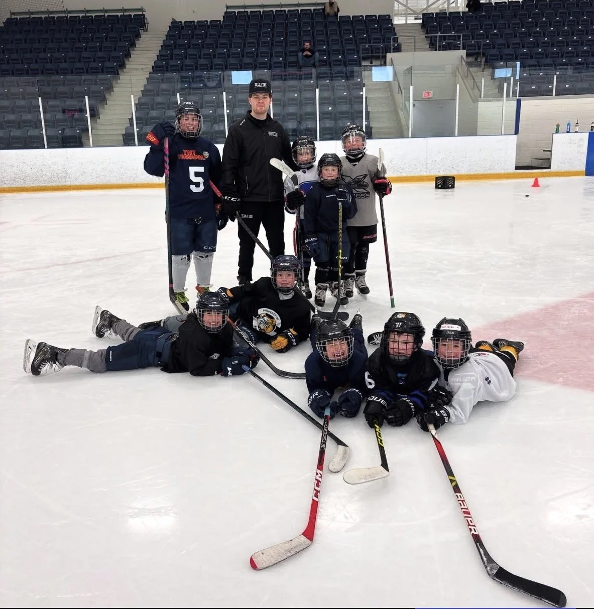 Group of children and a coach on an ice hockey rink, wearing hockey gear and helmets, posing for a team photo.