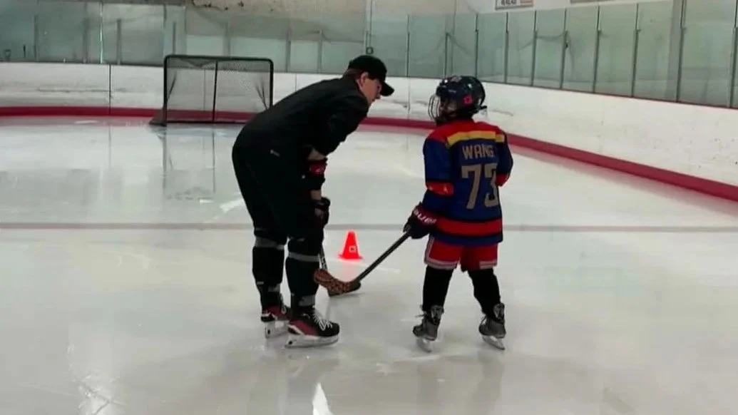 An adult hockey coach instructing a young child on ice hockey skills at an indoor ice rink, with a hockey goal and safety cone in the background.