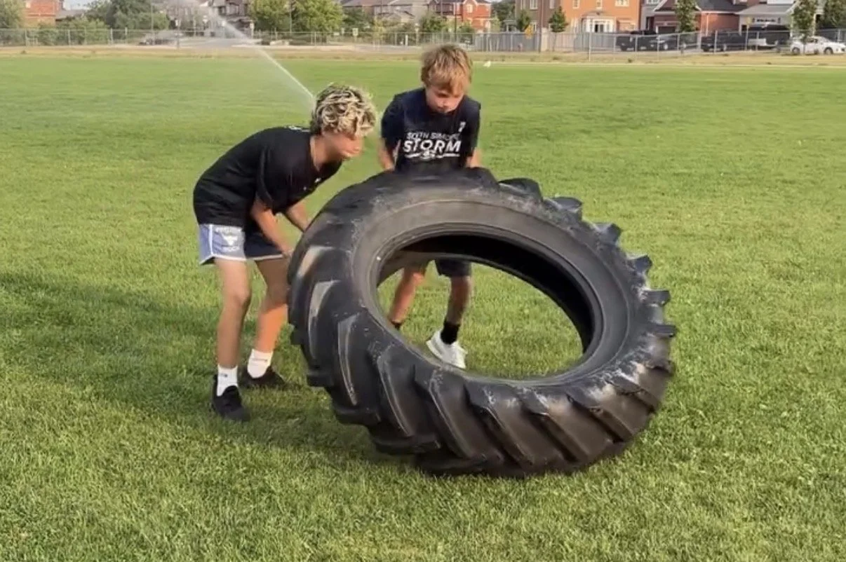 Two boys are playing with a large tractor tire on a grassy field, with houses and a fence in the background.